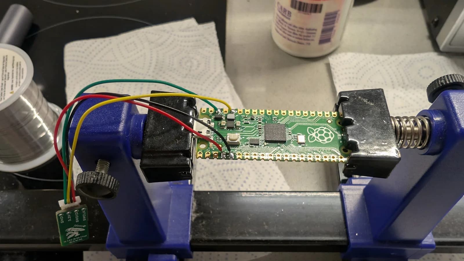 Raspberry Pi Pico (used as a debug probe) clamped in a blue PCB vise, coloured wires soldered to its pins running to a JST connector on a small WavePhoenix board at left; solder spool and supplies nearby.