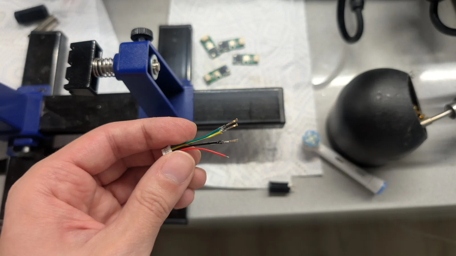 A hand holding a white JST connector whose coloured wires run off-frame; three of them have pins soldered on ready to fit the black cylindrical GameCube controller-port plug seen nearby, next to an electric toothbrush head. A blue soldering vise and small green WavePhoenix boards sit behind.