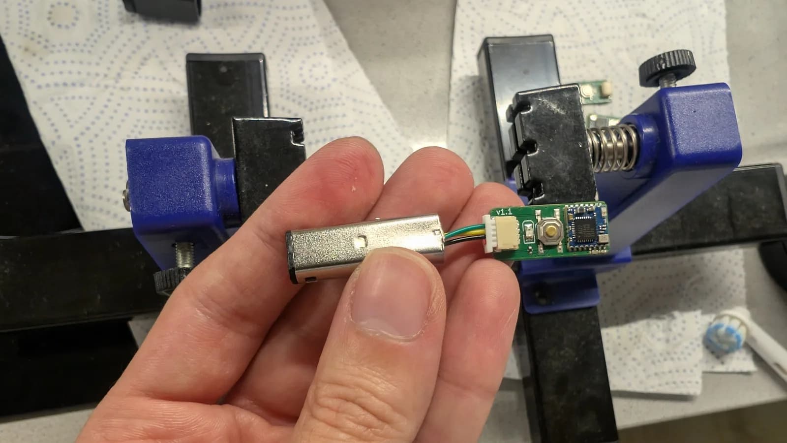 Hand holding a small green WavePhoenix board wired via a JST connector to a metal GameCube controller plug, a blue soldering vise behind.