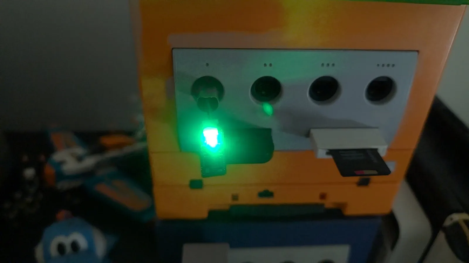 Small green WavePhoenix receiver plugged into port 1 of an orange GameCube, its green LED lit to show a paired WaveBird; the console's other controller ports and a memory card are alongside.