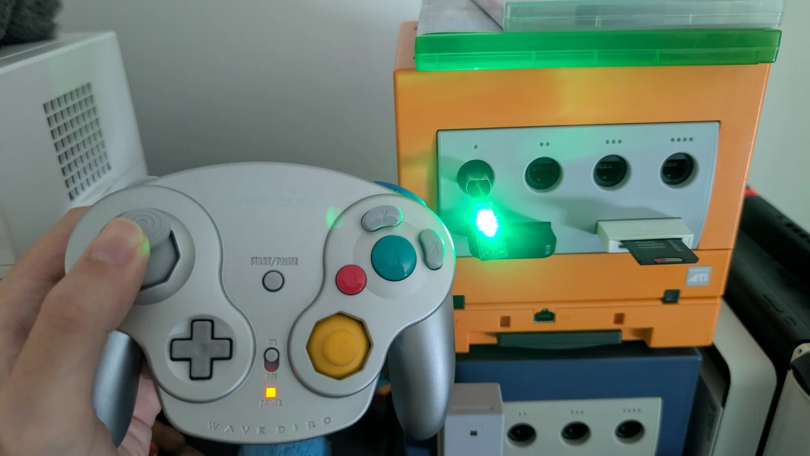 Hand holding a silver WaveBird wireless controller in front of an orange GameCube; the green WavePhoenix receiver in the console's port 1 has its green LED lit, showing the controller is paired.