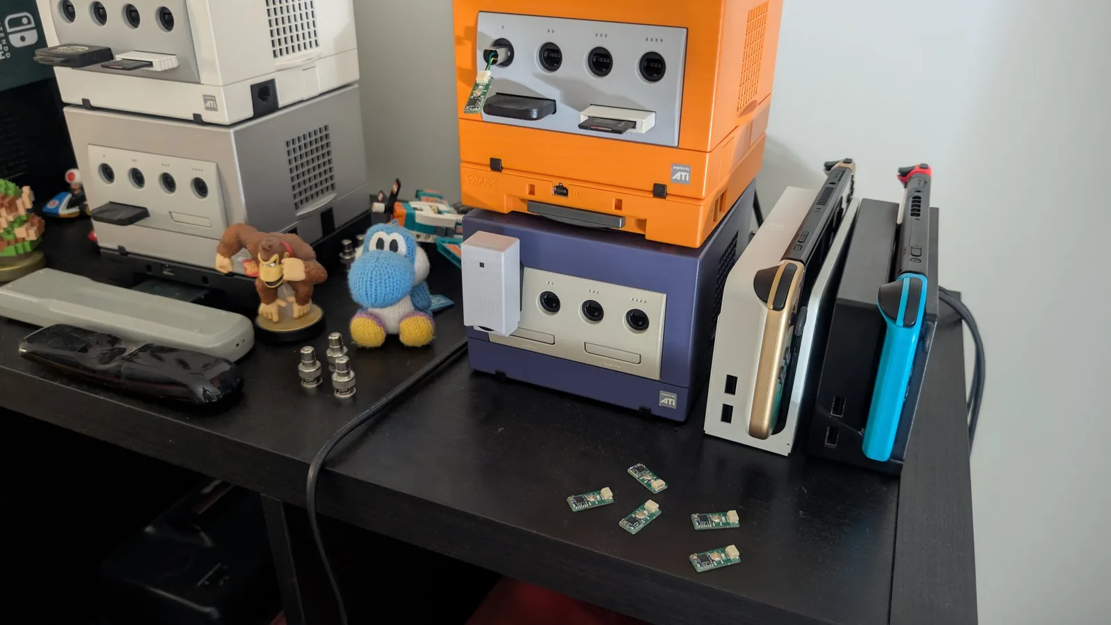Assembled WavePhoenix receiver boards on a black desk being tested in front of a collection of GameCube consoles and Nintendo Switch units, with one receiver plugged into the orange GameCube's port 1.