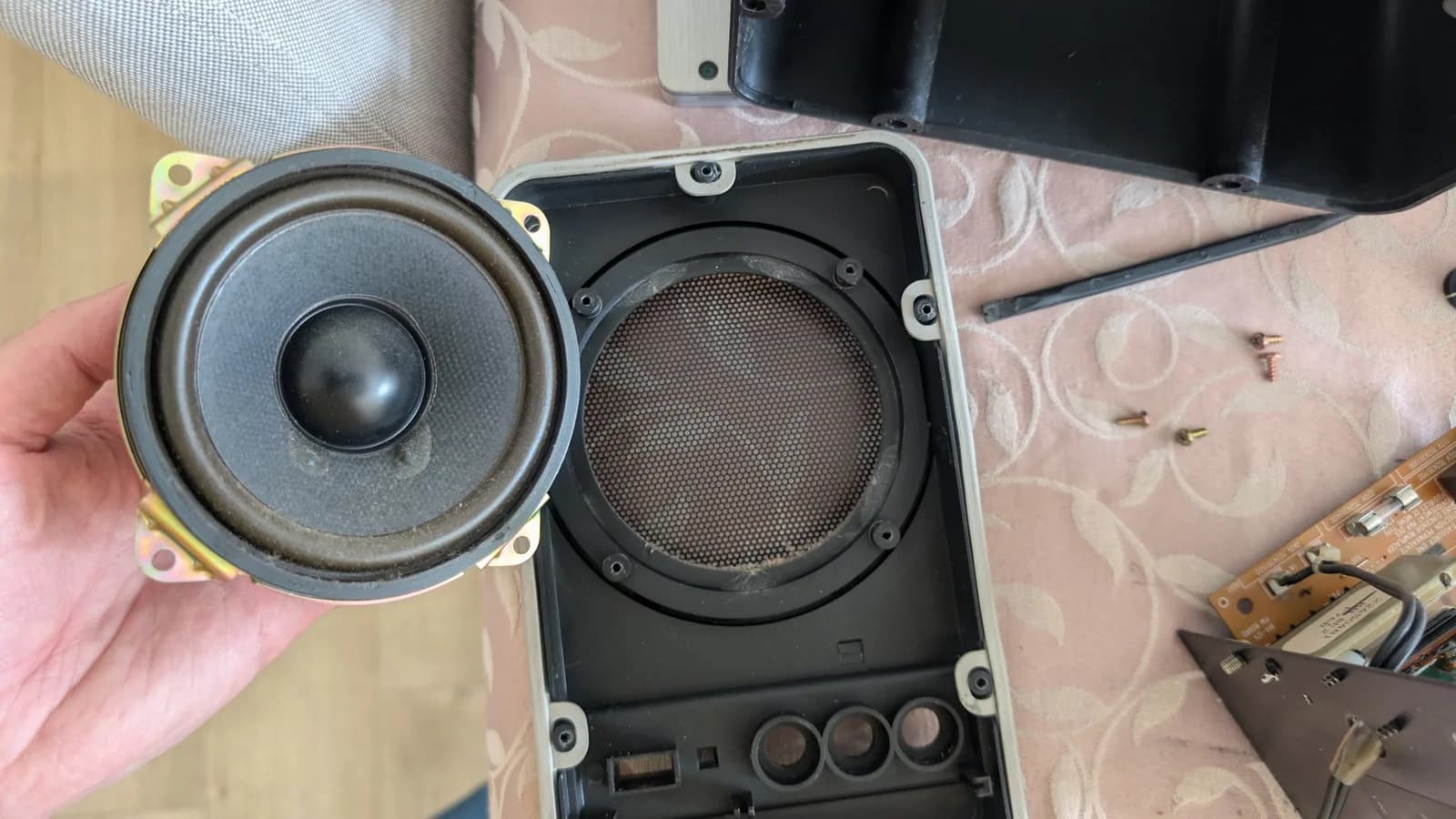 Disassembled BOSS MA-12AV speaker before cleaning, its driver removed and held in hand, the dusty black cone and surround facing the camera; the front baffle with its grimy internal grille mesh alongside.