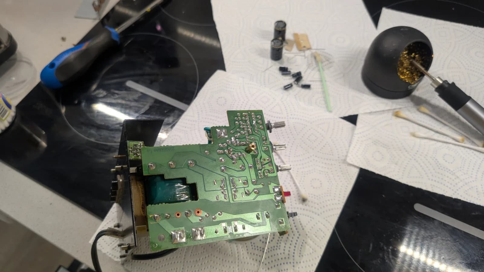 Solder side of a BOSS MA-12AV amplifier board, its joints freshly reflowed and shiny, a soldering iron resting in its brass-coil tip cleaner to the side; the removed old capacitors on the paper towel behind.