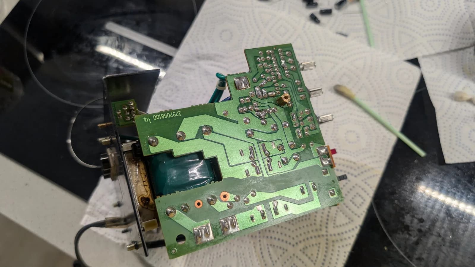 Close-up of the solder side of a BOSS MA-12AV amplifier board after a full reflow, the joints freshly shiny, with the pot shafts and a small transformer visible; a couple of removed old capacitors and a swab on the paper towel behind.