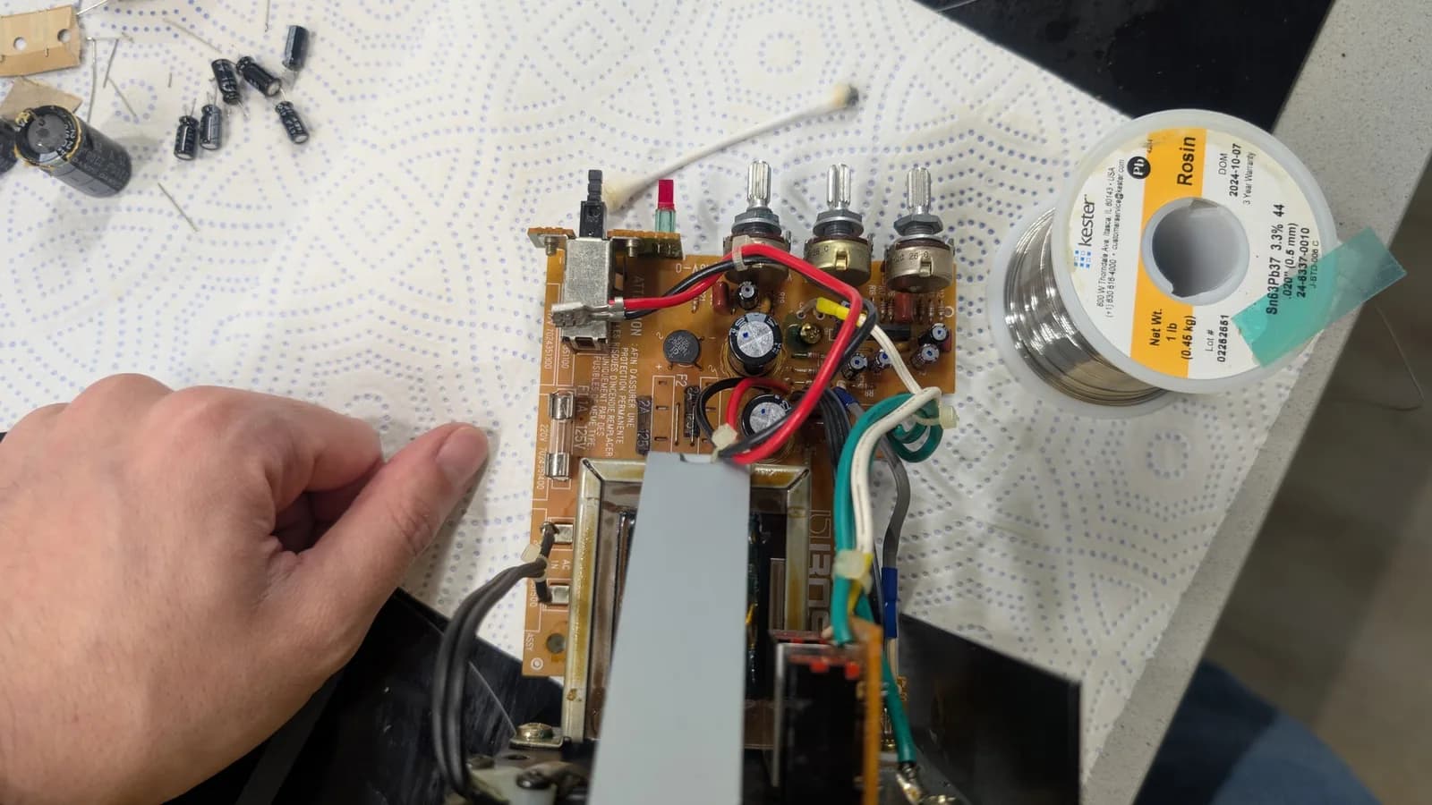 Top view of a recapped BOSS MA-12AV amplifier board with its new capacitors fitted and each marked with a dot, the removed old capacitors off to the side; a solder spool and a cotton swab alongside.