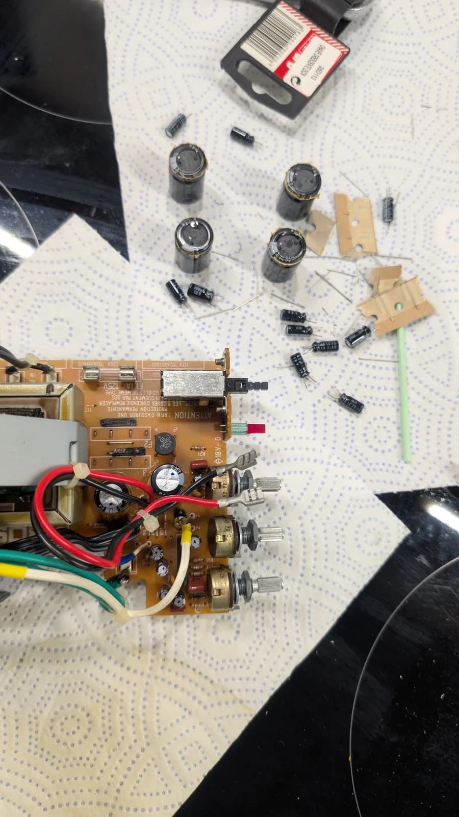 Top view of a recapped BOSS MA-12AV amplifier board with its new capacitors fitted and marked with a dot, and the removed old capacitors from both units in shot on the blue-dotted paper towel.
