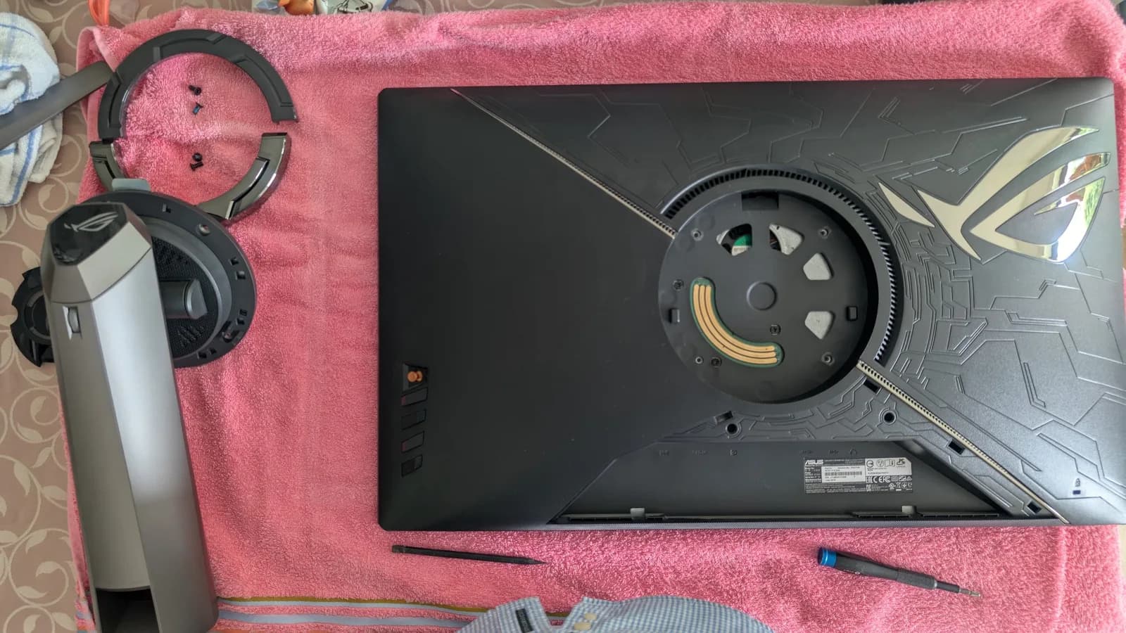Asus PG27UQ monitor bottom panel removed showing internal fan assembly with gold connectors and black shroud during refurbishment, headphones and screwdriver visible.
