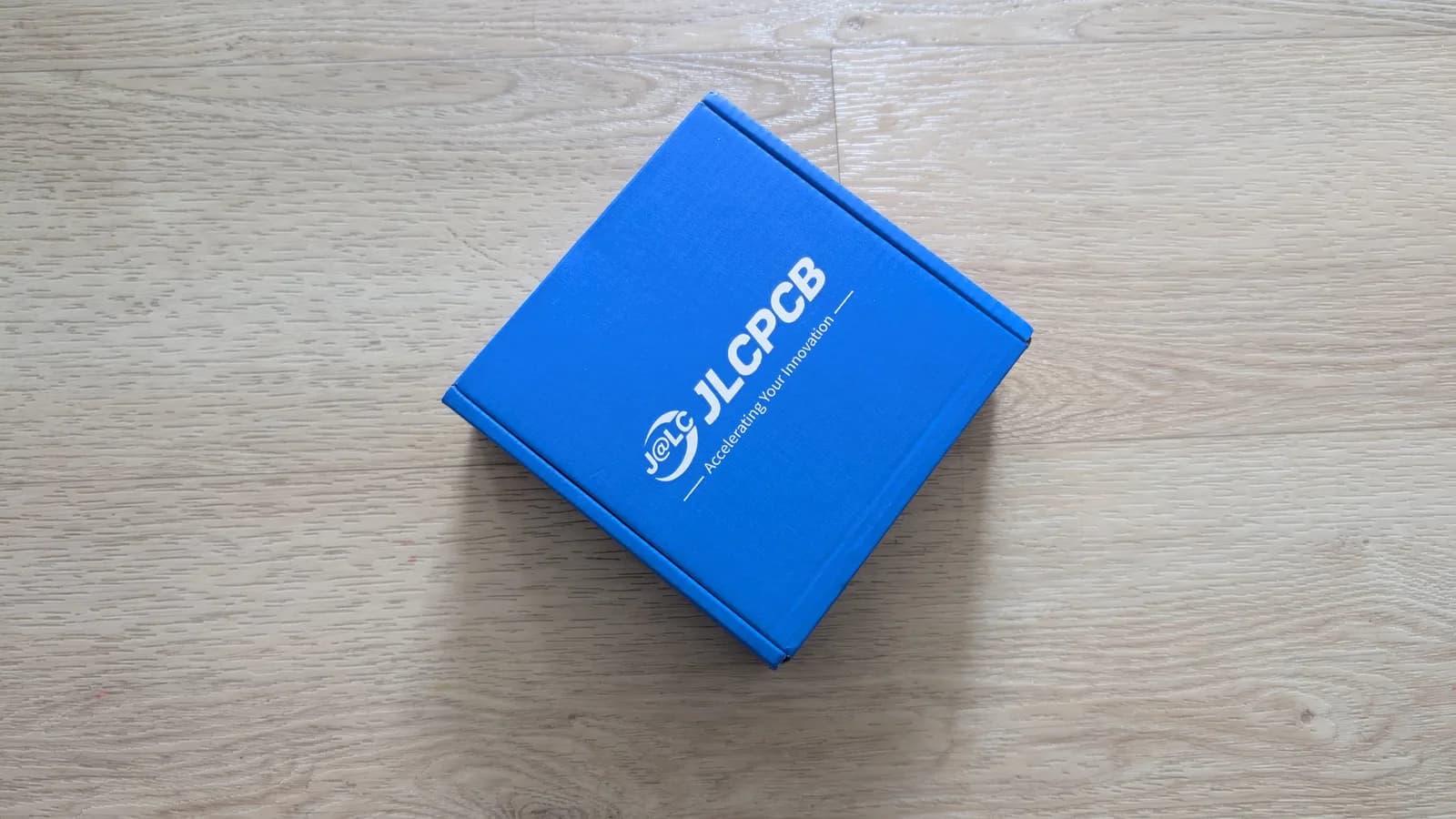 Closed blue JLCPCB shipping box on a light wood surface, the delivery of pre-assembled WavePhoenix receiver boards.
