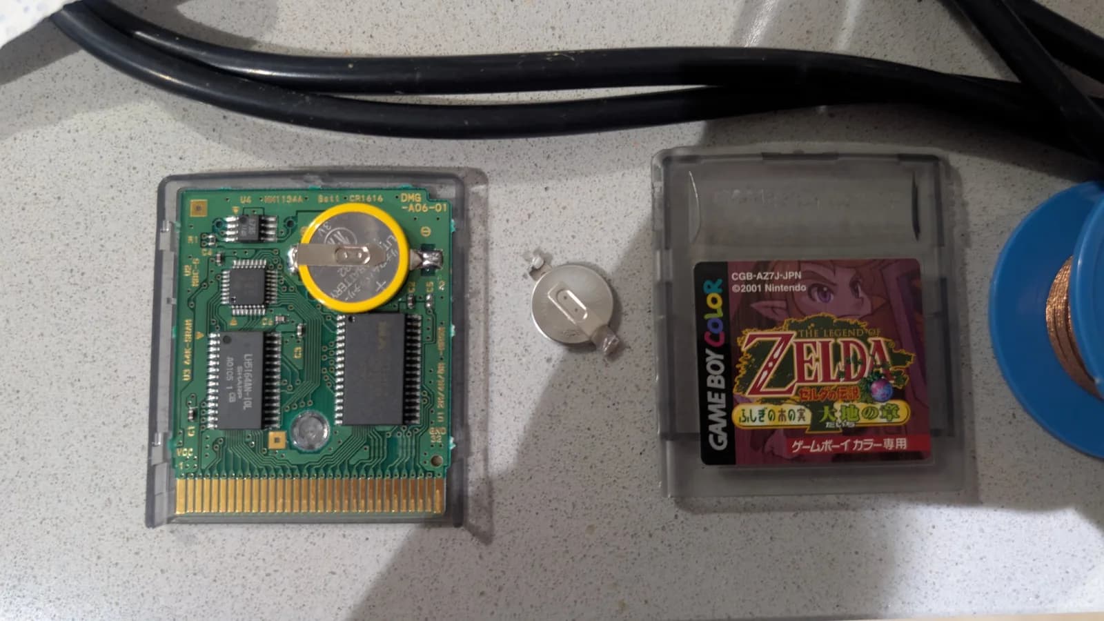 Two Japanese Game Boy Color cartridges mid save-battery swap. The left cartridge is open, its green PCB showing a fresh tabbed CR2032 coin cell (its edge wrapped in yellow plastic) soldered in place, with the removed old tabbed cell resting beside it. The right is The Legend of Zelda: Oracle of Seasons (CGB-AZ7J-JPN) in its clear shell, and a blue spool of desoldering braid sits at the edge of frame.