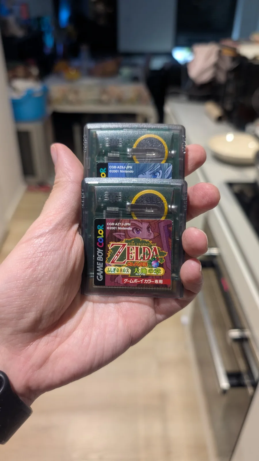 A hand holding two clear-shell Japanese Game Boy Color cartridges stacked together: in front, The Legend of Zelda: Oracle of Seasons (CGB-AZ7J-JPN); behind it, The Legend of Zelda: Oracle of Ages (CGB-AZ8J-JPN). The yellow ring of each cartridge's save battery is visible through the translucent shell.