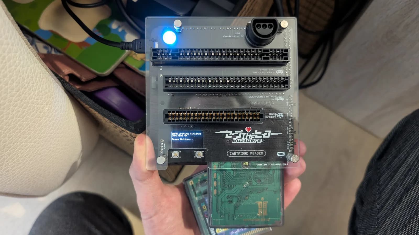 A Save The Hero Builders cartridge reader (an Open Source Cartridge Reader build) held in hand, its blue power LED lit. The board has slots for SNES/SFC, Sega Genesis/MD, N64 and GBA/GB-GBC carts plus an N64 controller port, and its small OLED reads 'SRAM writing finished, Verified OK'. A Game Boy cartridge is seated in the bottom GB/GBC slot.