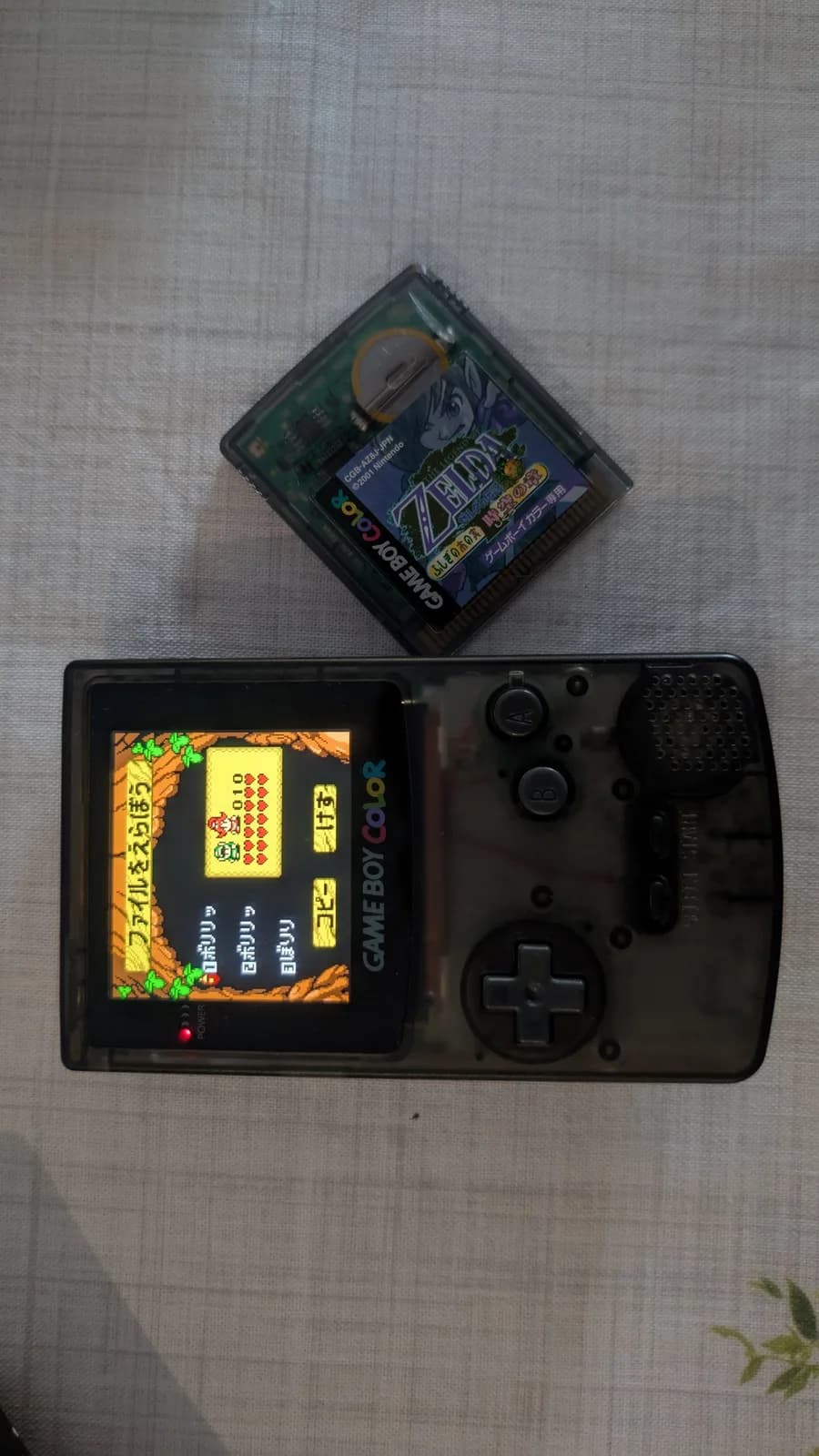 A translucent-black Game Boy Color powered on, showing the file-select screen of The Legend of Zelda: Oracle of Seasons (the cartridge running in it), with the other game, The Legend of Zelda: Oracle of Ages (CGB-AZ8J-JPN), resting on the cloth just above it.