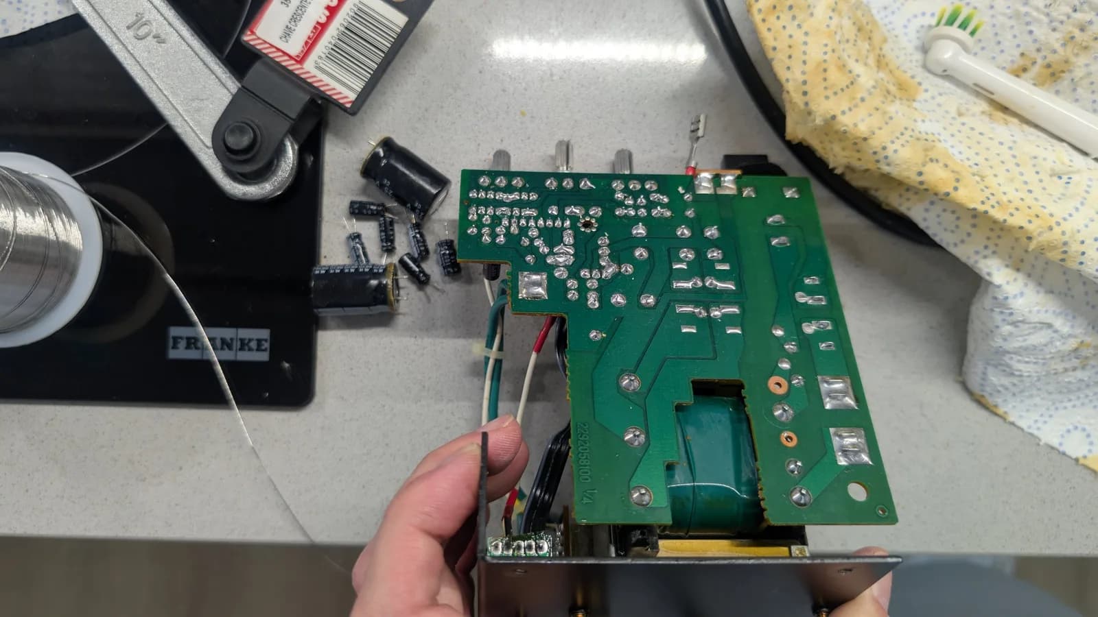 Hands holding the green BOSS MA-12AV amplifier board (solder side) during the recap, all its joints freshly reflowed and shiny; the removed old capacitors on the work surface.