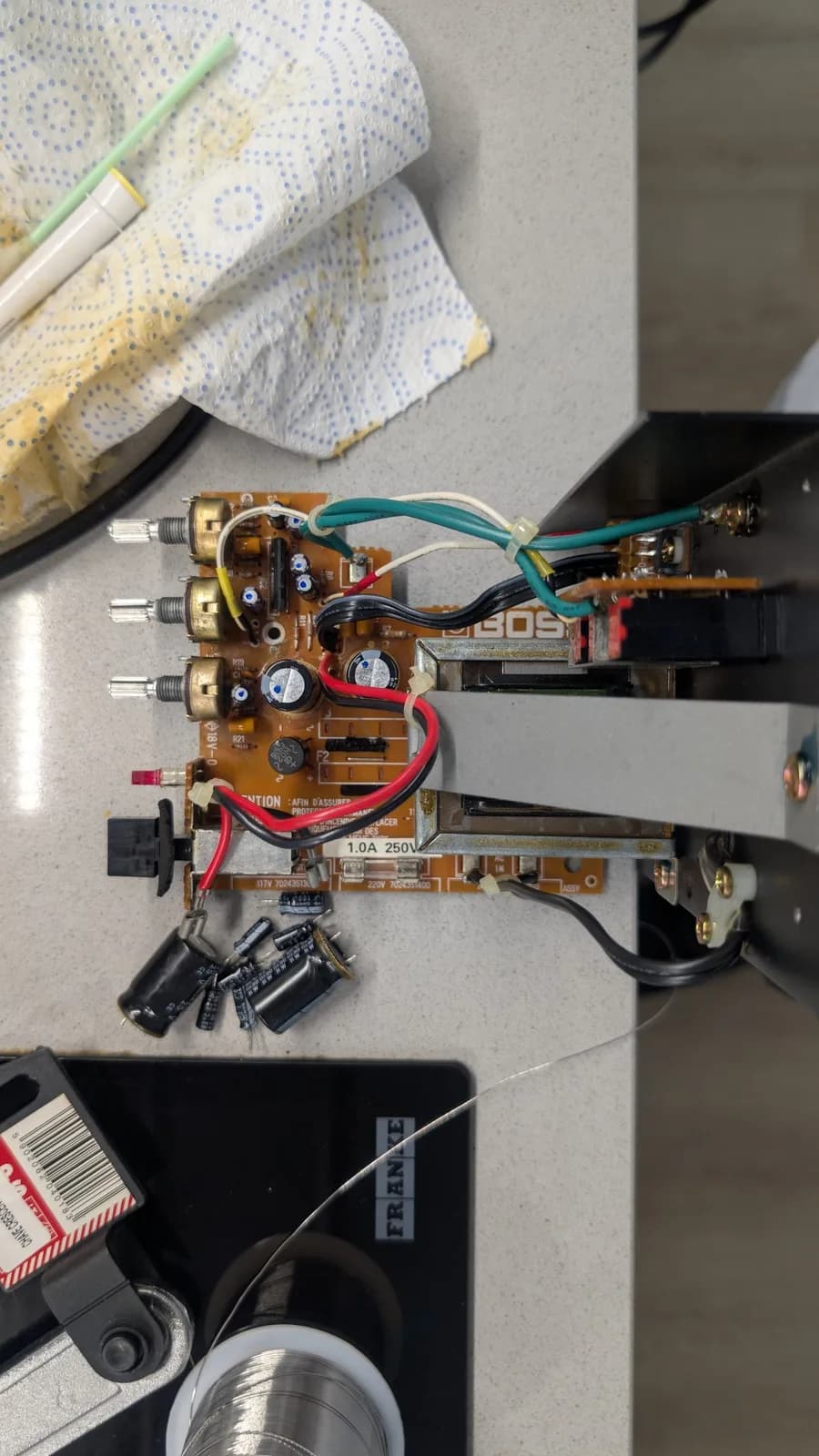 Top view of the recapped BOSS MA-12AV amplifier board with its new capacitors fitted and each marked with a dot, the removed old capacitors alongside the PCB; wiring, a blue-dotted paper towel and a brush head above.