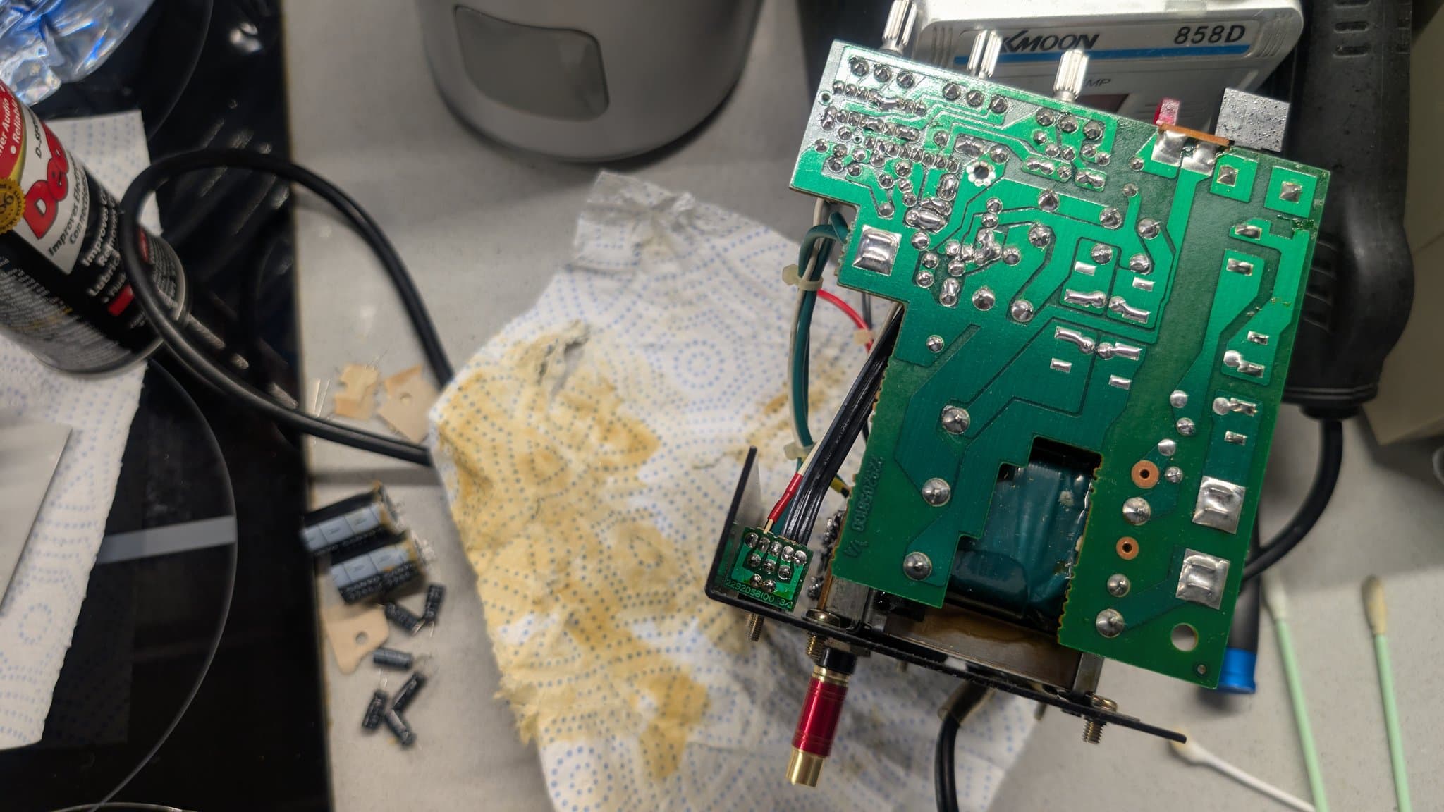 Green PCB with exposed capacitors and components during recapping work; yellow dotted cloth and desoldering tools visible on workbench.