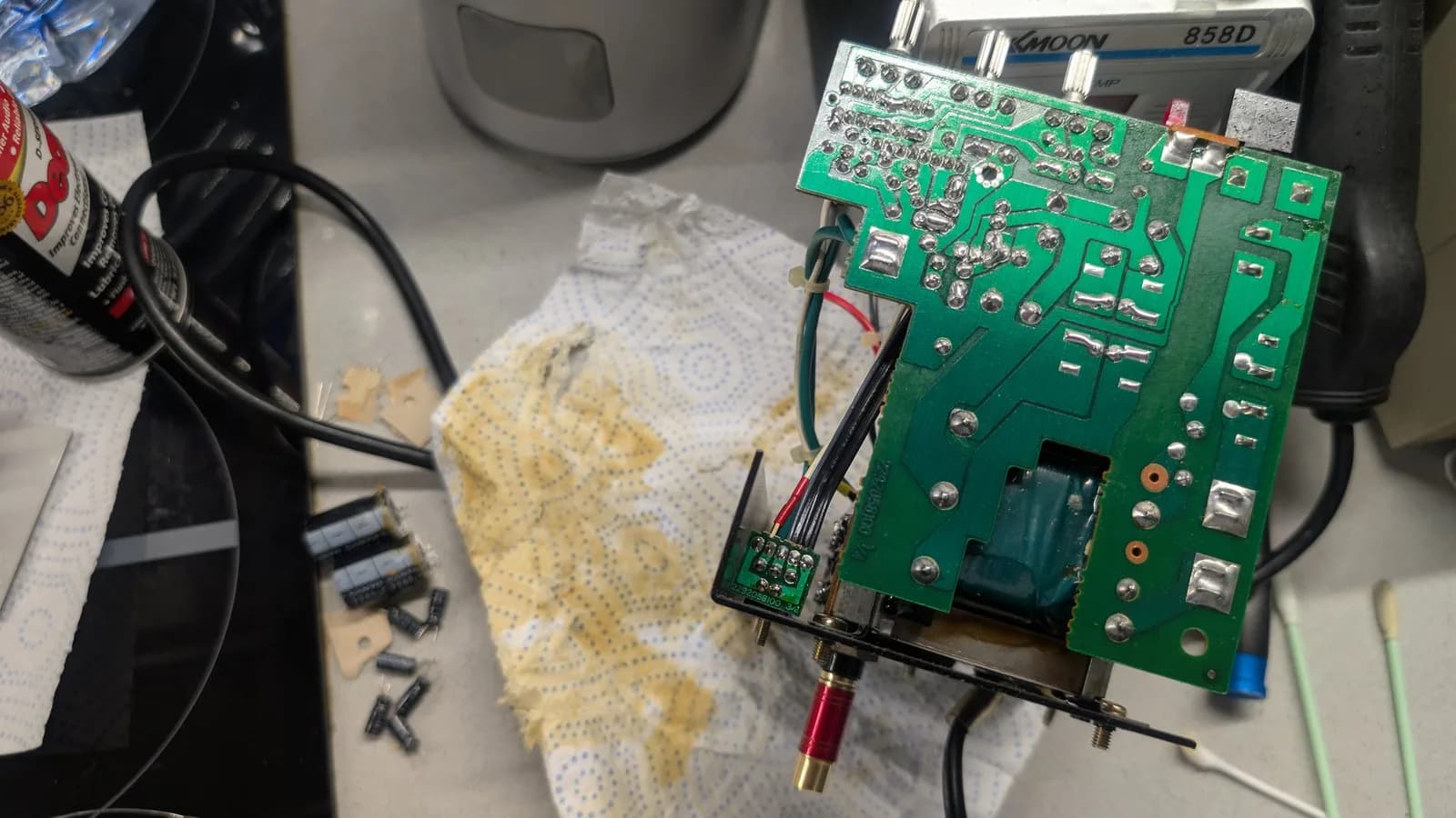 Solder (trace) side of the second speaker unit's BOSS MA-12AV amplifier board, its joints freshly reflowed and shiny; removed old capacitors, cotton swabs and a can of contact cleaner on the workbench beside a blue-dotted paper towel, a hot-air rework station behind.