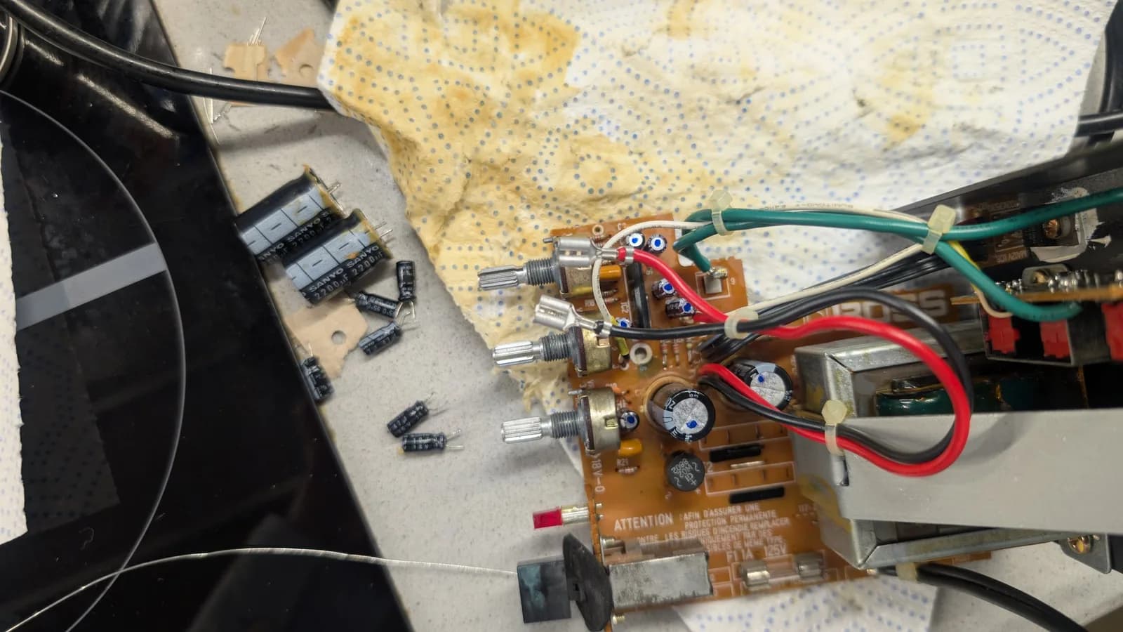 Top view of the second speaker unit's BOSS MA-12AV amplifier board with its new capacitors fitted and marked with a dot, and the removed old electrolytic capacitors (including two large Sanyo 2200uF cans) off to the side.