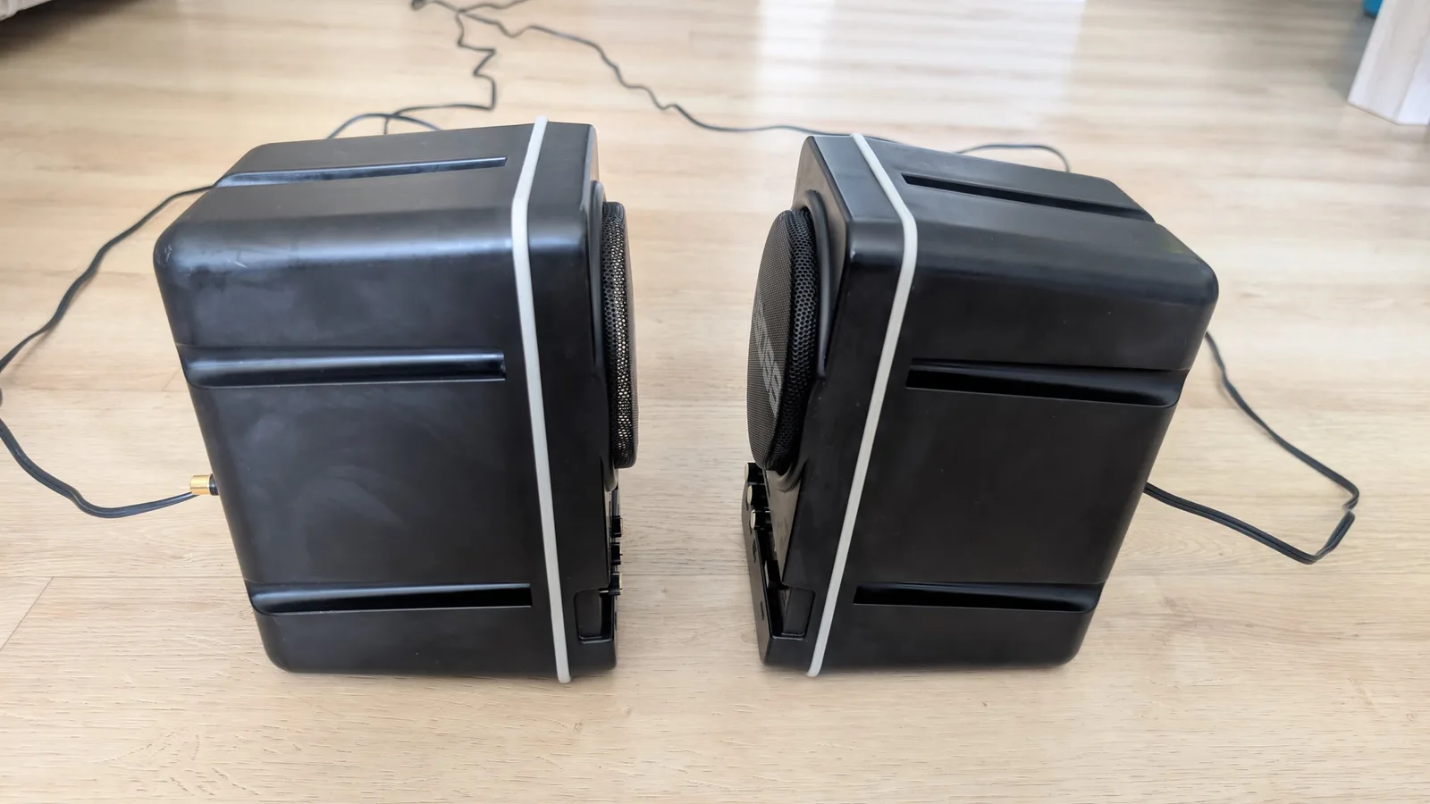 Side view of the pair of refurbished BOSS MA-12AV speakers on a wooden floor, their cleaned black plastic enclosures shown from the side, with a cable running across.