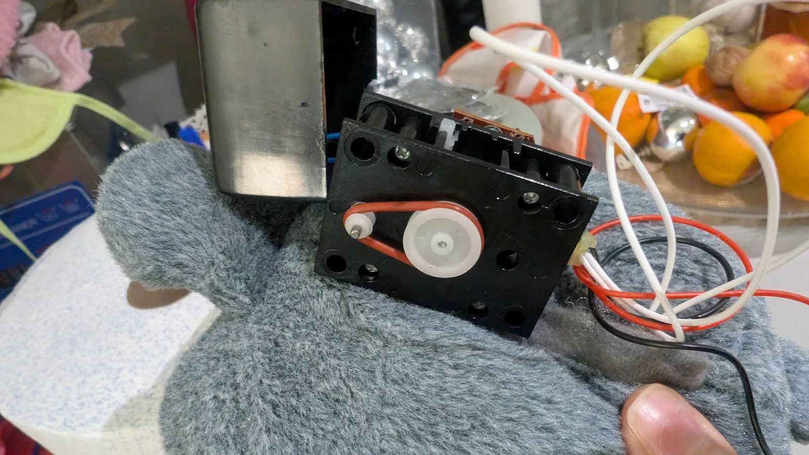 Close-up of Totoro toy mechanism showing black plastic gear assembly with white and orange gears, foam padding, and red/white wiring on gray fabric surface.