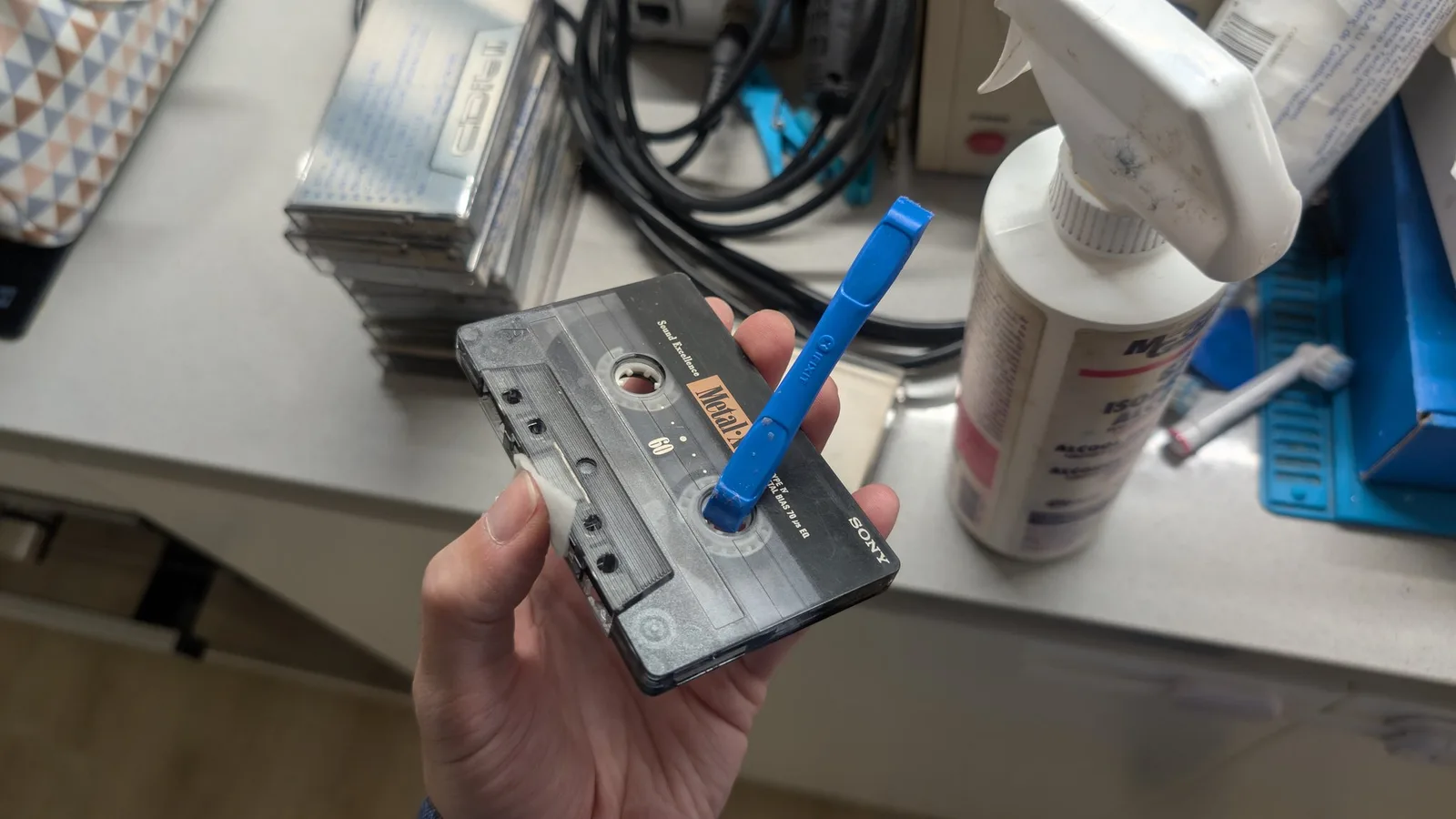 Hand holding a Sony cassette tape with blue pen inserted in reel, cleaning supplies and cables visible on workbench.
