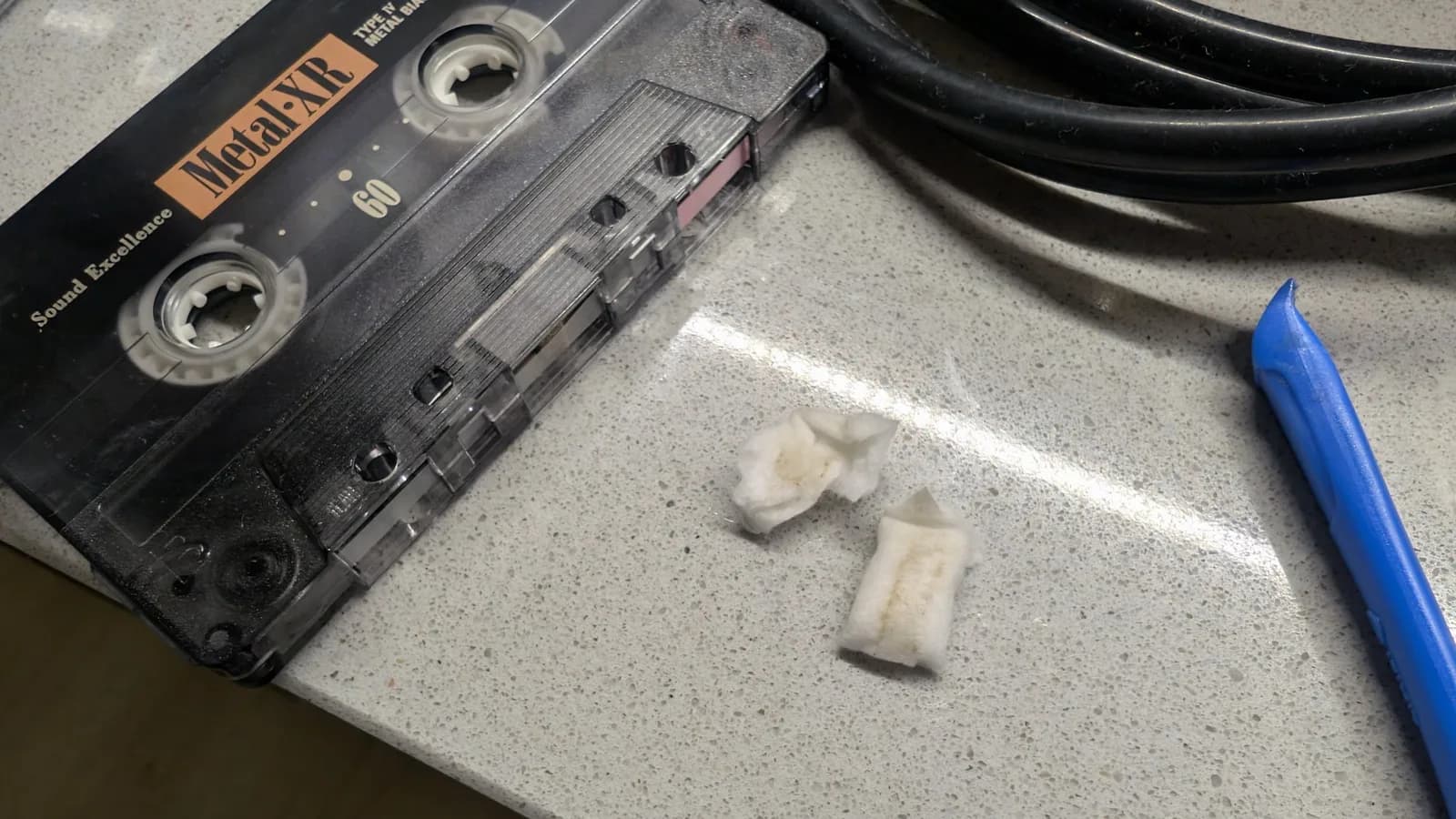 Maxell cassette tape with spooled tape beside blue rewinding tool on work surface during cleaning restoration.