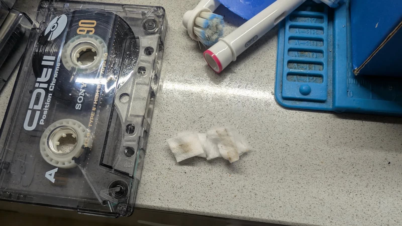 Cassette tape maintenance setup: Akai Type II cassette with cleaning supplies, cotton swabs, and blue tape deck on workbench surface.