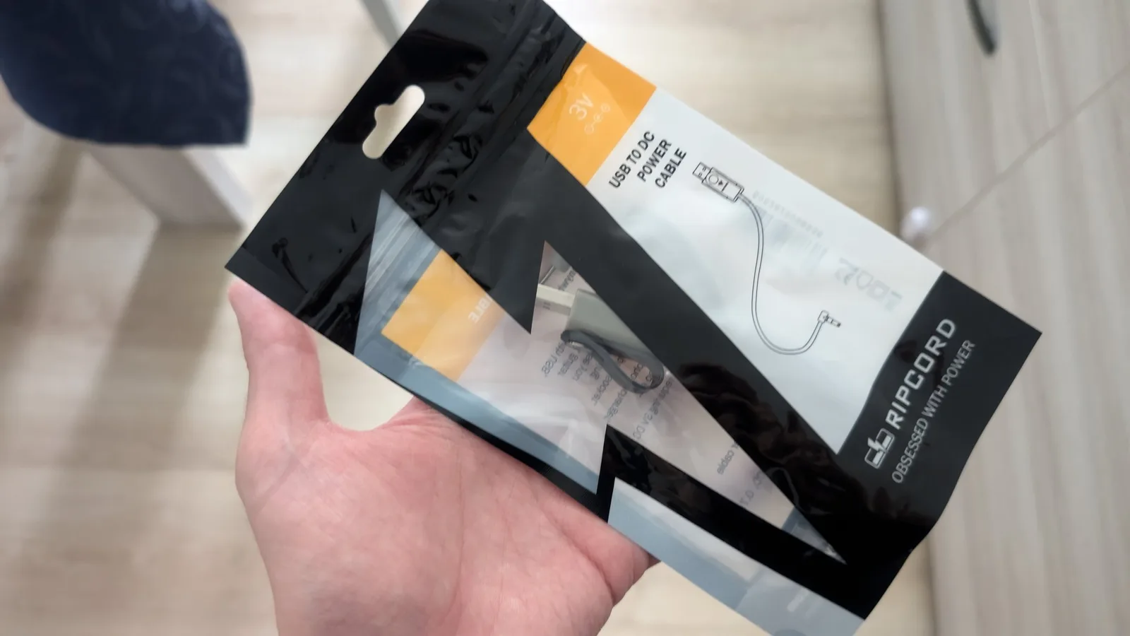 Hand holding MiniDisc charging cable package with USB power adapter and cord visible through clear packaging window.