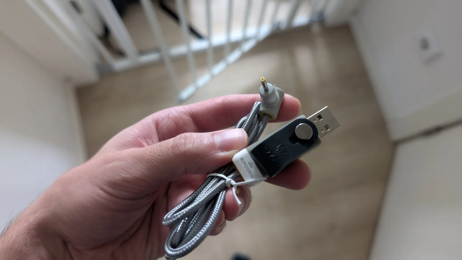 Hand holding a USB charging cable with braided gray sleeve connected to a MiniDisc player charger adapter.