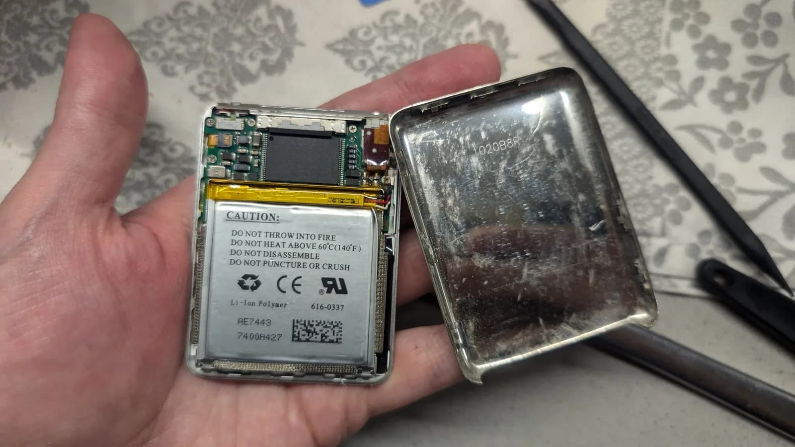 Disassembled iPod Nano showing Li-Ion polymer battery, circuit board, and yellow connector. Caution label visible on battery casing.