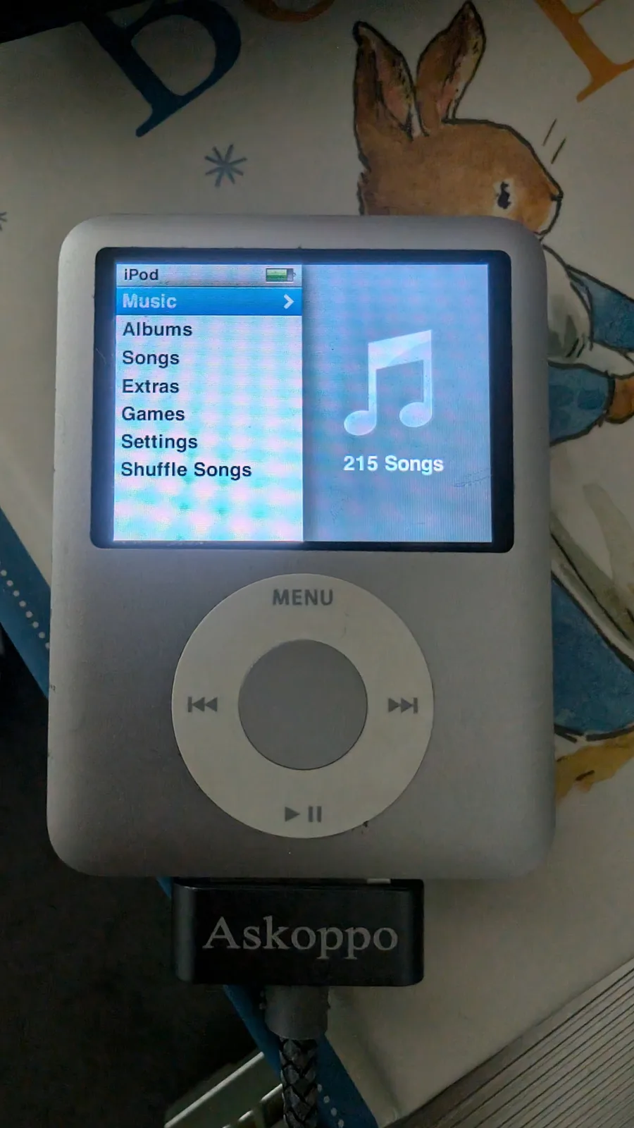 White iPod Nano displaying Music menu with 215 songs, connected to Askoppo charger dock, on repair workbench with rabbit illustration visible.