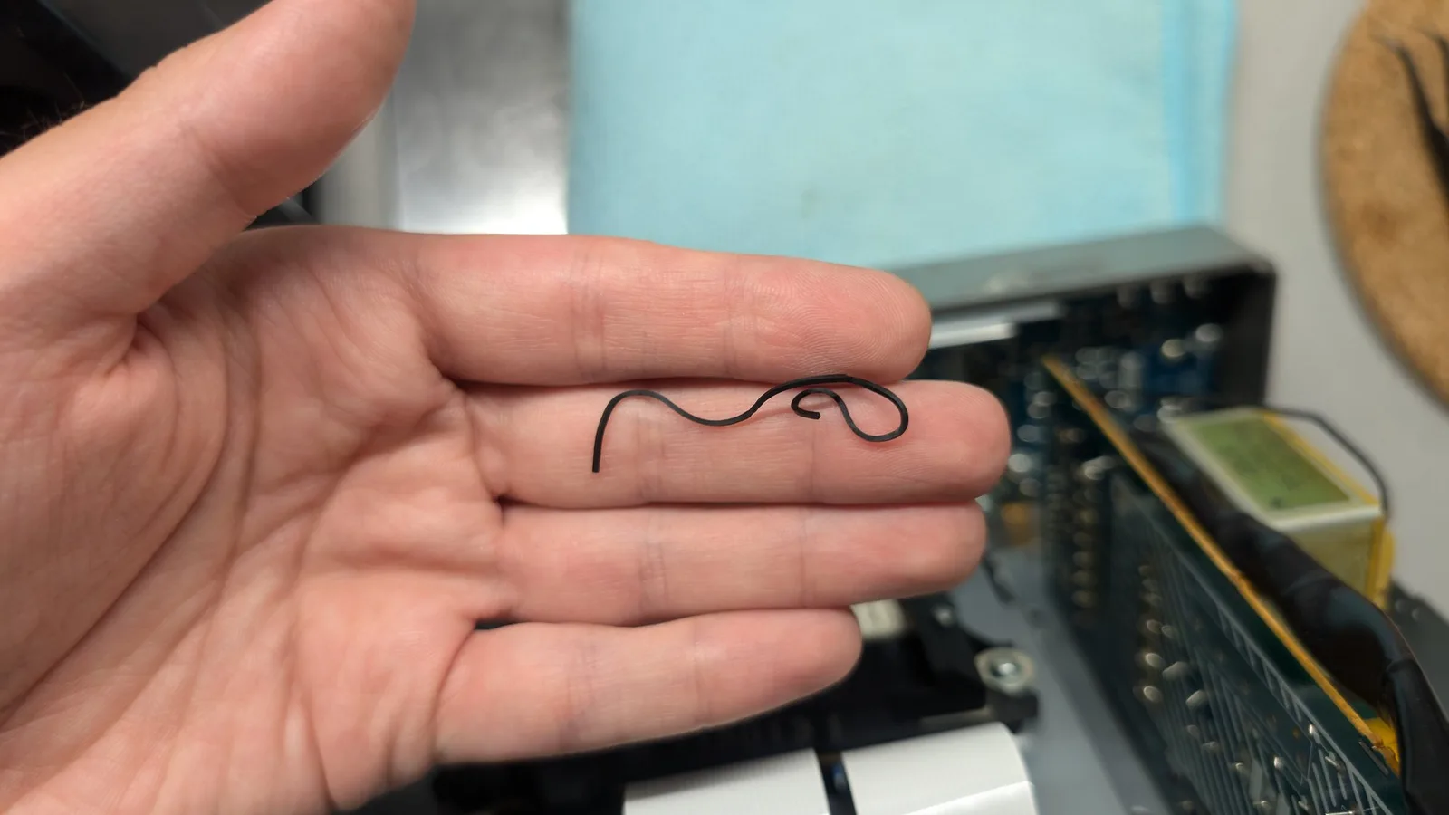 Hand holding a worn rubber belt shaped like a coiled snake against a workbench with electronic equipment in the background.