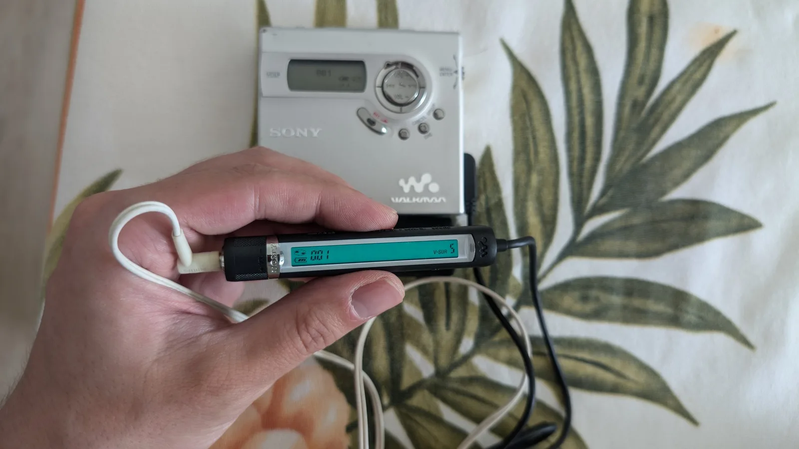 Hand holding Sony MiniDisc remote controller with turquoise LCD display showing 88.1 FM, MZ-N920 portable player visible in background.