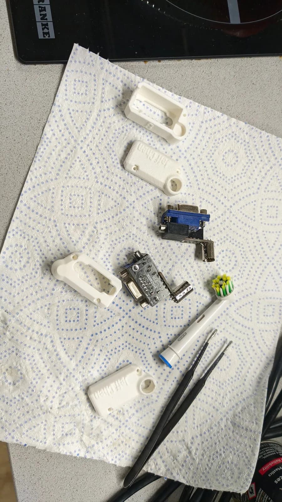 Disassembled 8DIN2VGA (Famicom) adapter parts on a paper towel: white 3D-printed shell halves (embossed 'Jeff Chen'), the assembled boards with blue VGA connectors, metal connectors and yellow SMD components, plus an electric toothbrush head and tweezers.