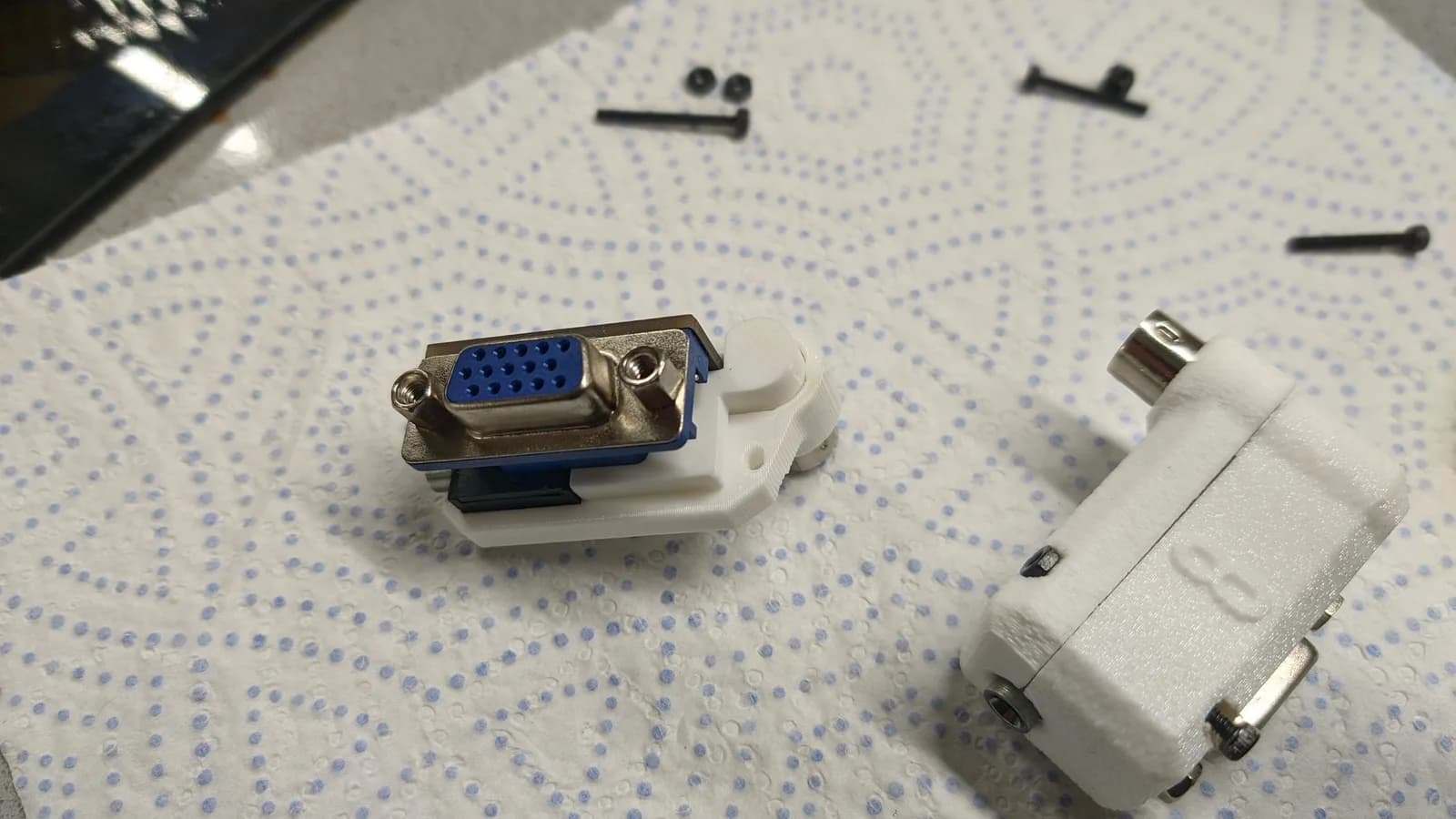 Two white-shelled 8DIN2VGA (Famicom) dongles on a dotted paper towel, showing how they assemble into the shell: one is still open, revealing a white support piece fitted inside the shell that holds the dongle, and its blue VGA connector; the other is closed, showing the metal 8-pin DIN (Famicom-side) connector; mounting screws sit above.