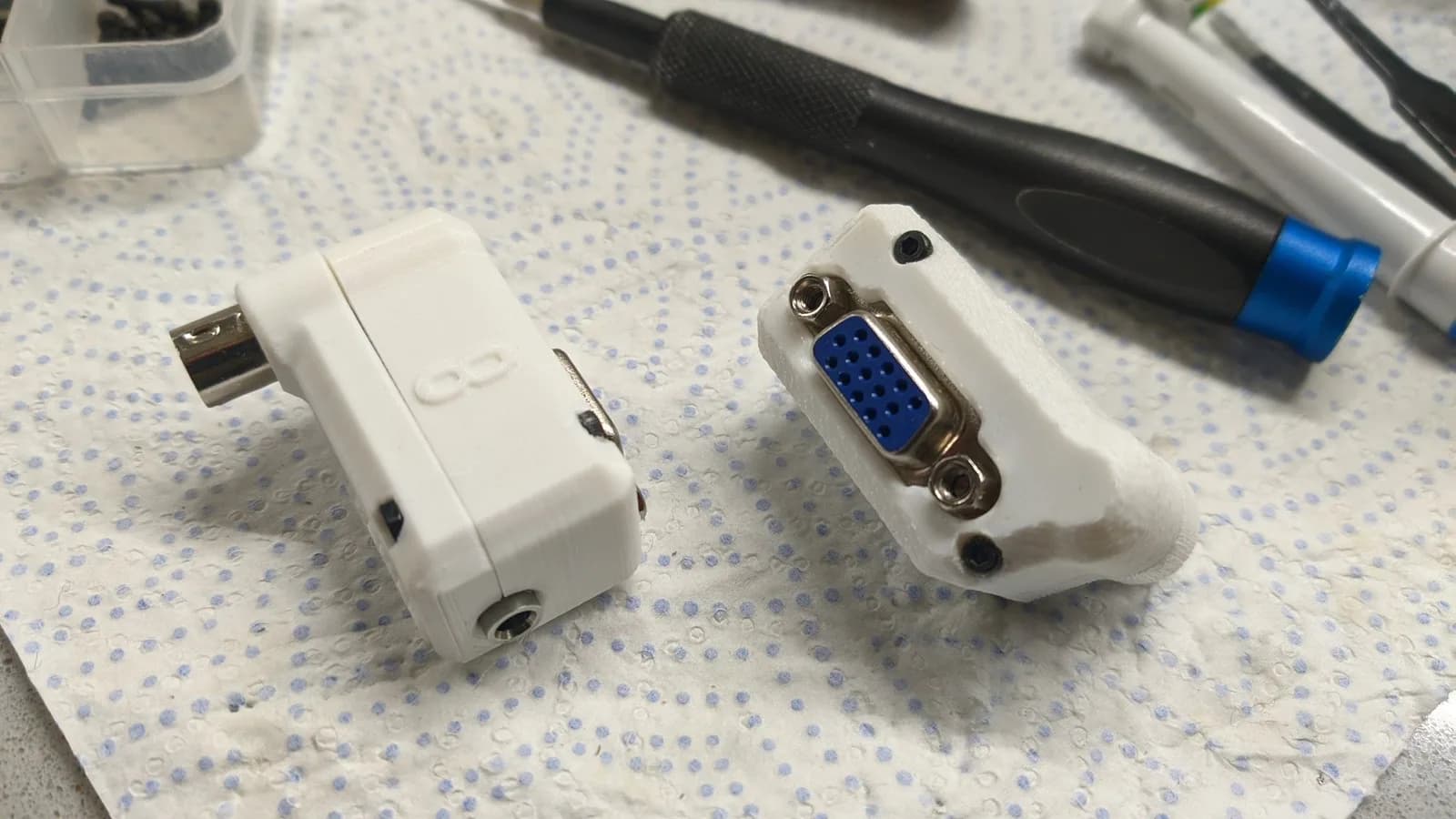 Two white-shelled 8DIN2VGA (Famicom) dongles on a dotted paper towel: the left showing its metal 8-pin DIN (Famicom-side) connector and a 3.5mm jack, the right showing its blue VGA connector; a precision screwdriver and an electric toothbrush lie behind.