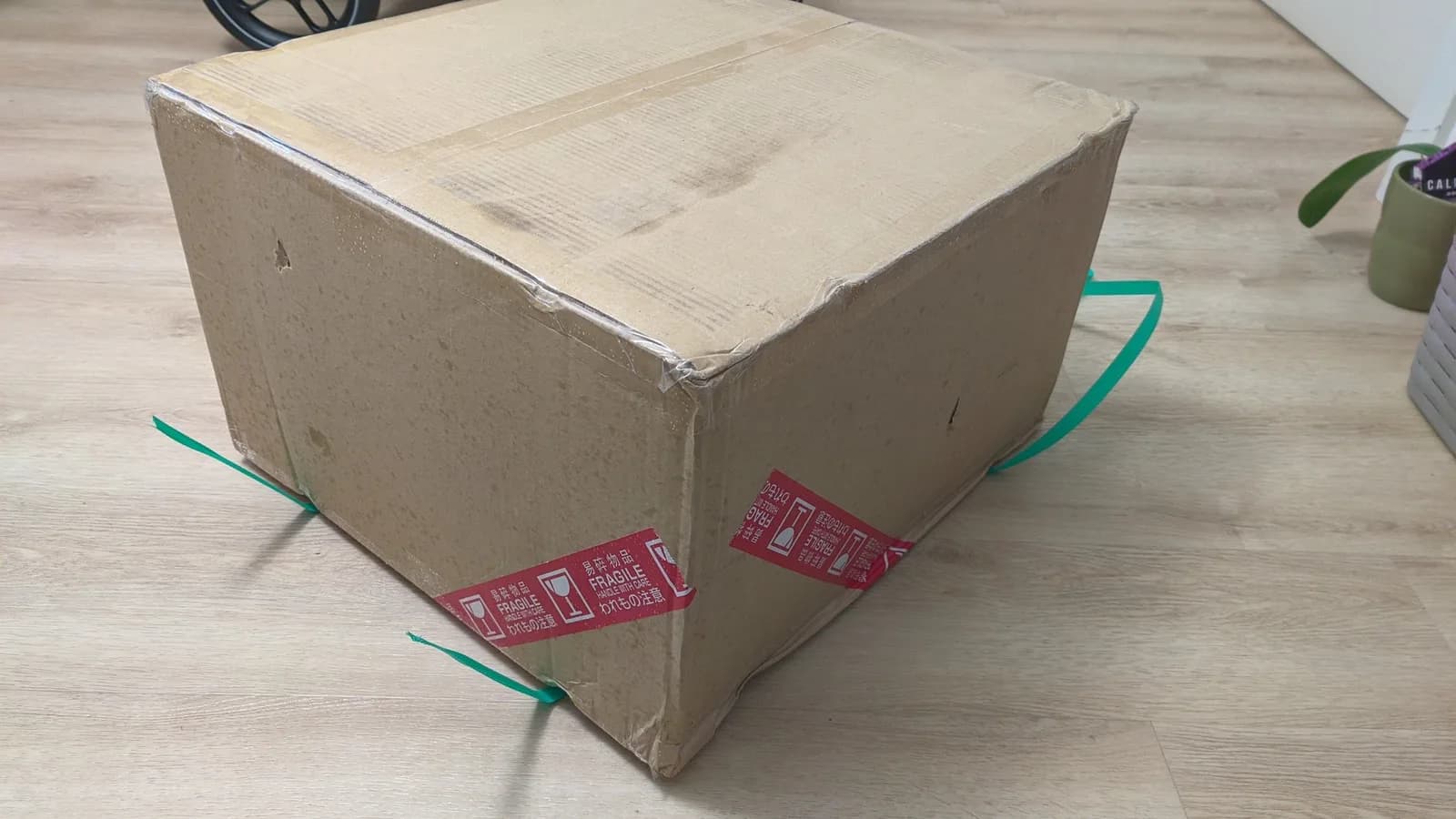 Cardboard shipping box with green twine handles and red Sendico tape, showing insufficient protective packaging for fragile CRT monitor shipment.