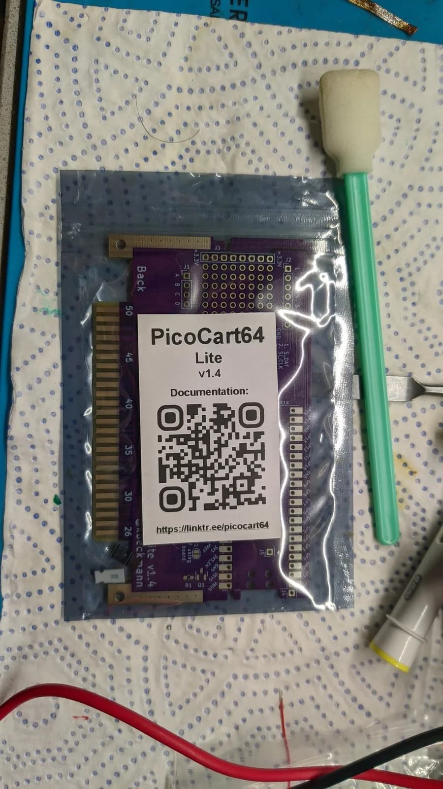 PicoCart64 Lite v1.4 cartridge in protective bag with foam brush and documentation QR code visible on workbench.