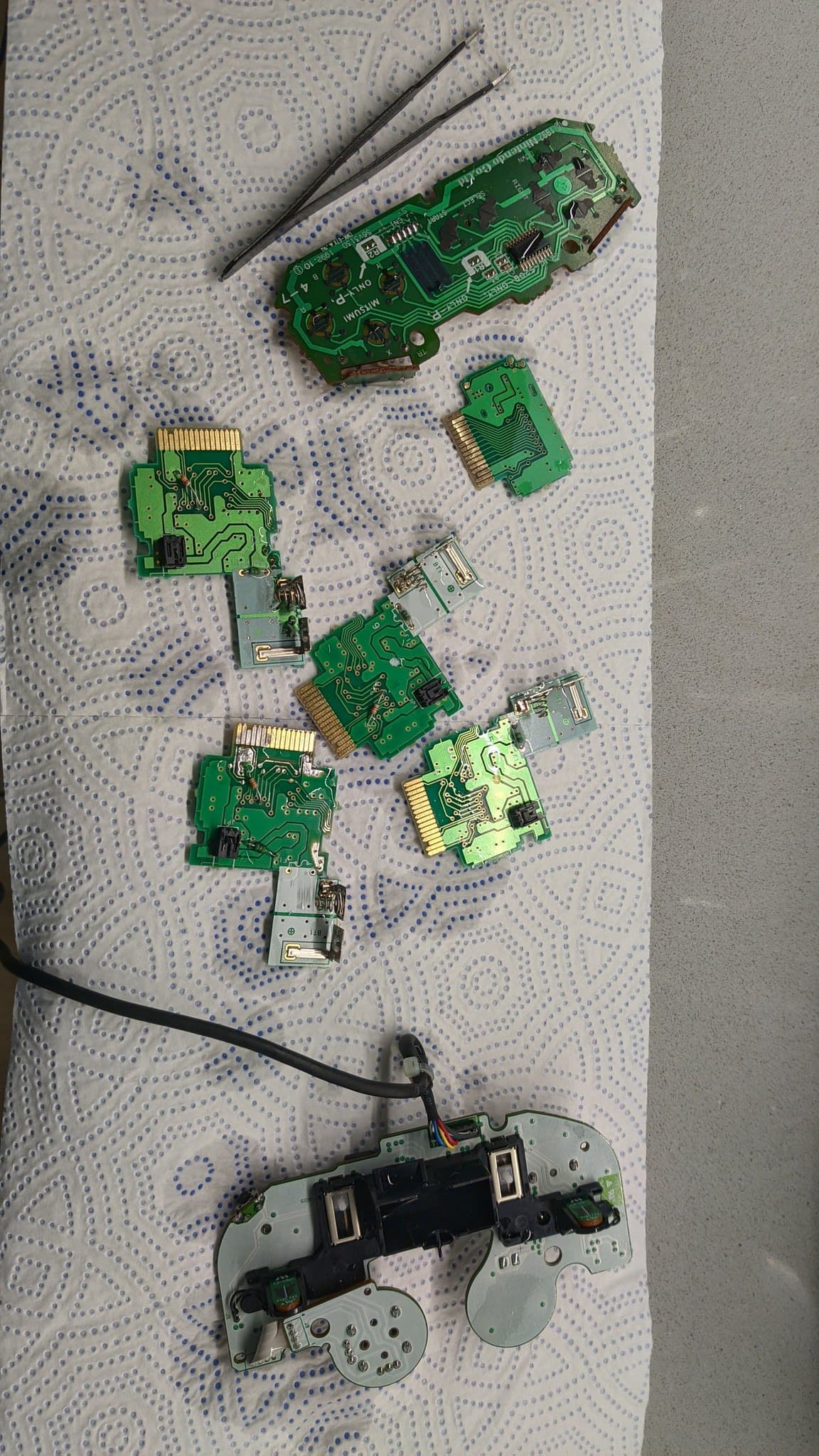 Disassembled controller PCBs and rumble pack boards laid out on patterned fabric, cleaned via ultrasonic cleaner with tweezers nearby.