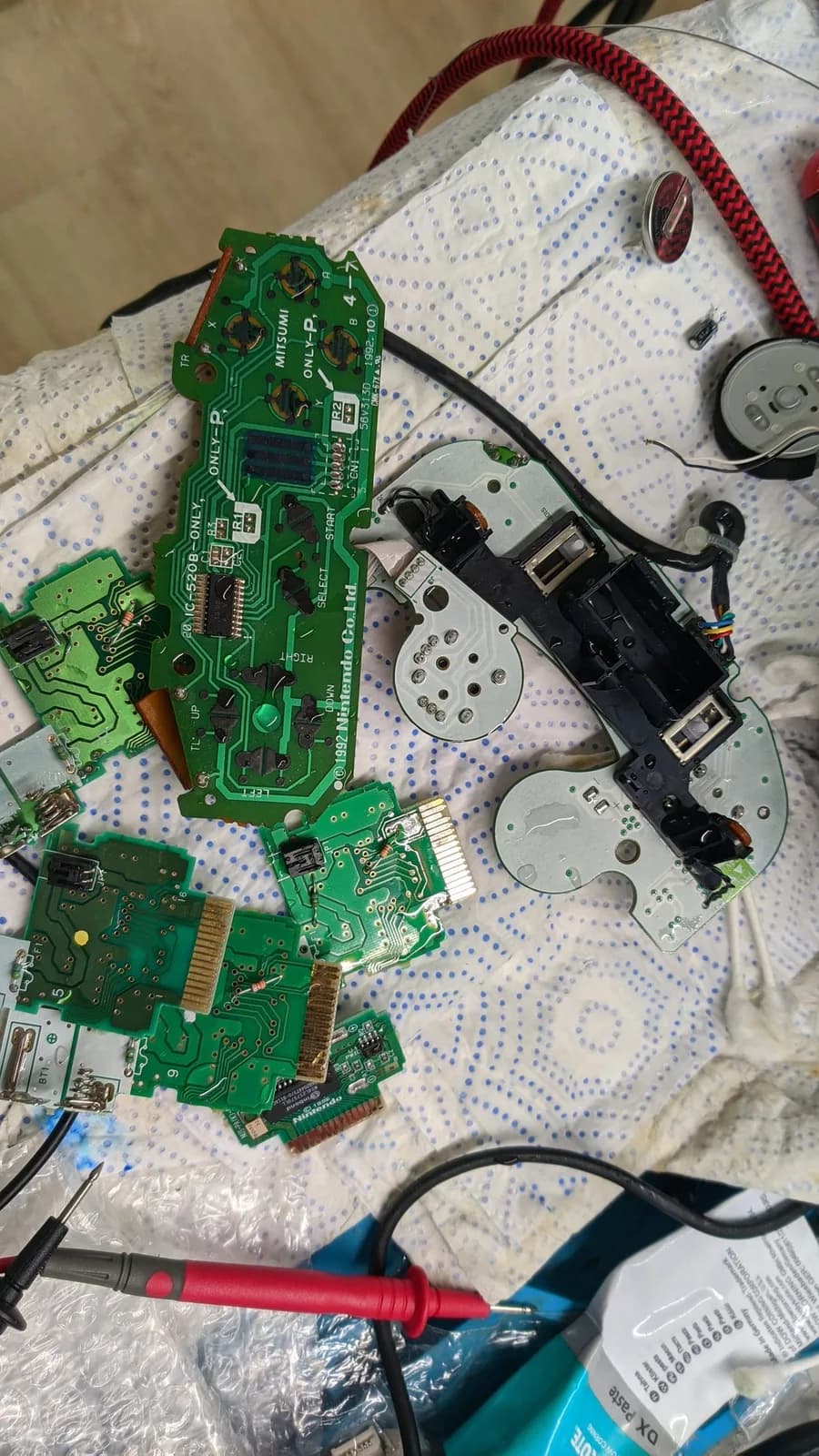 Disassembled controller rumble packs and PCBs on breadboard after ultrasonic cleaning, with multimeter probe visible.