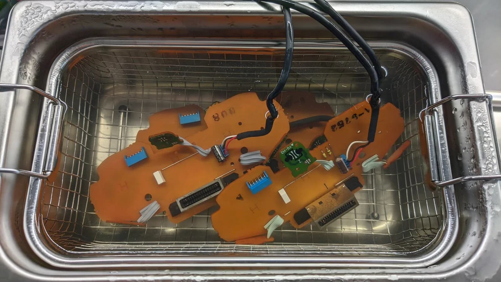 Orange PCBs and controller shells in ultrasonic cleaner basket with soapy water, showing capacitors, connectors, and circuit traces after cleaning.