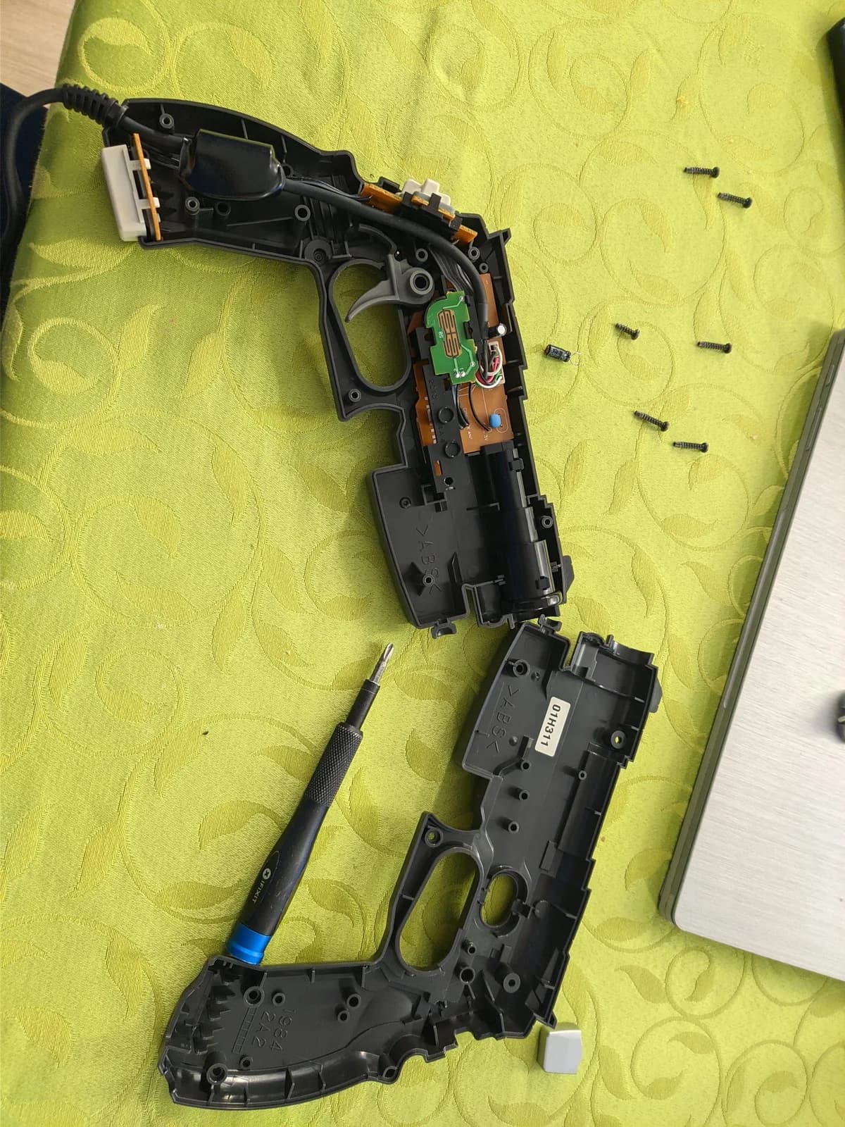 Disassembled GunCon and GunCon 2 light guns showing internal PCBs, green capacitors, and screwdriver on yellow work mat during repair.