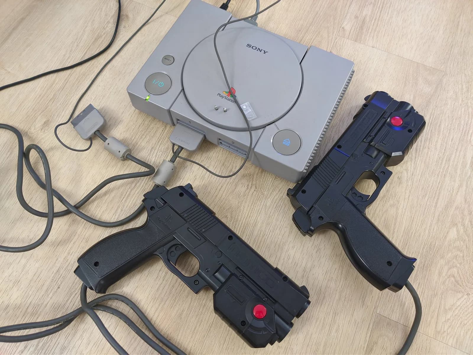 Two GunCon light guns with red trigger buttons connected to original PlayStation console via cables on wooden surface.