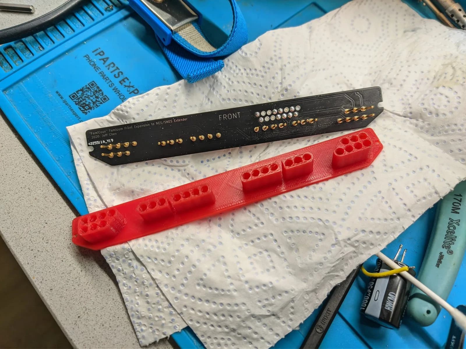 Black NES controller extension board with gold connectors next to red 3D-printed adapter, both on white work mat.