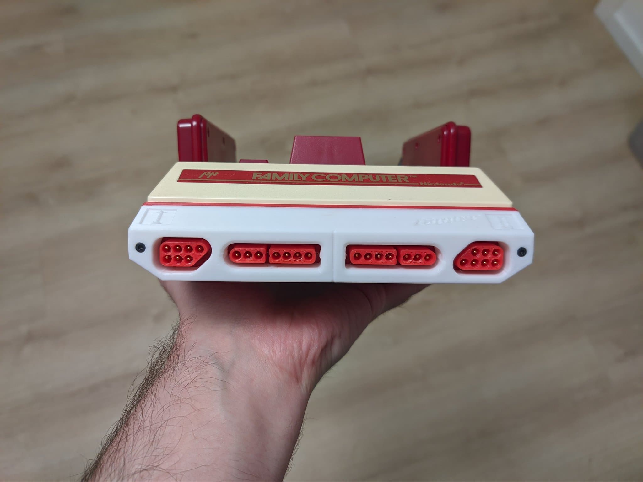 Hand holding Famicoun NES controller adapter with white body, red connectors, and red controller ports on top.