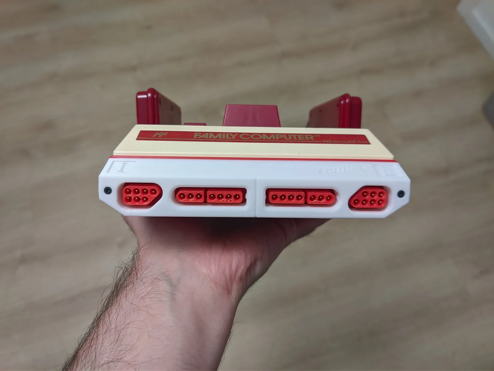 Hand holding Famicoun NES controller adapter with white body, red connectors, and red controller ports on top.