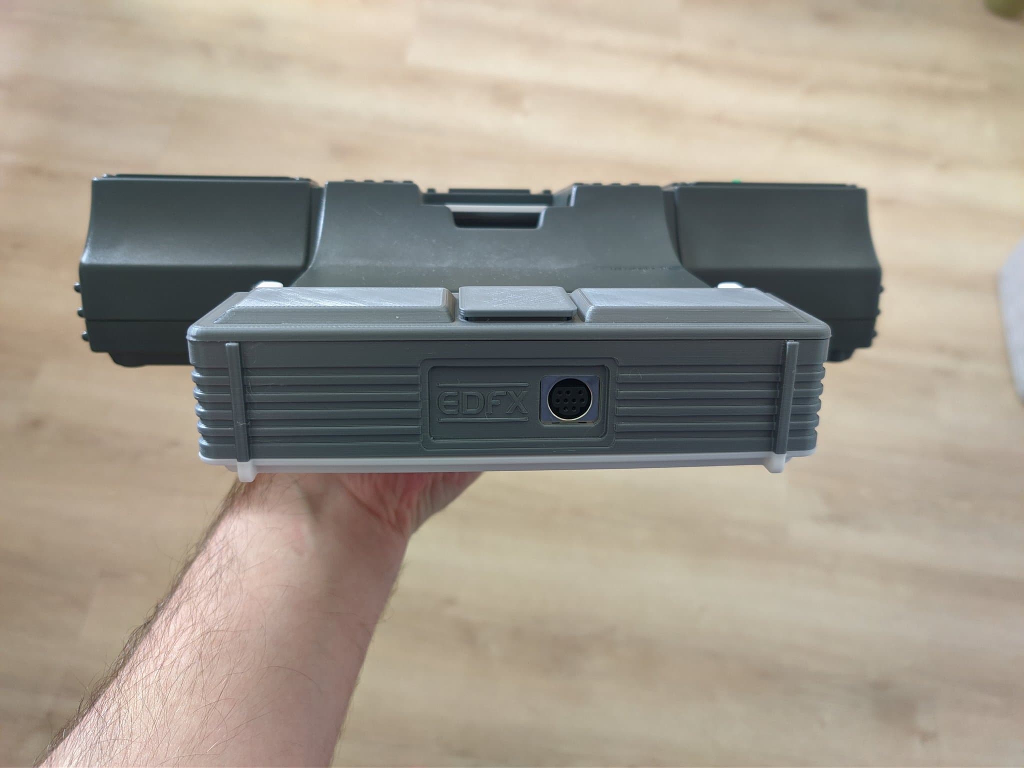 Front view of the Krikzz EDFX adapter in its 3D-printed mini-IFU shell, held in hand above a wooden surface, showing the EDFX branding and its 9-pin DIN output port, which takes a Mega Drive 2 RGB cable.