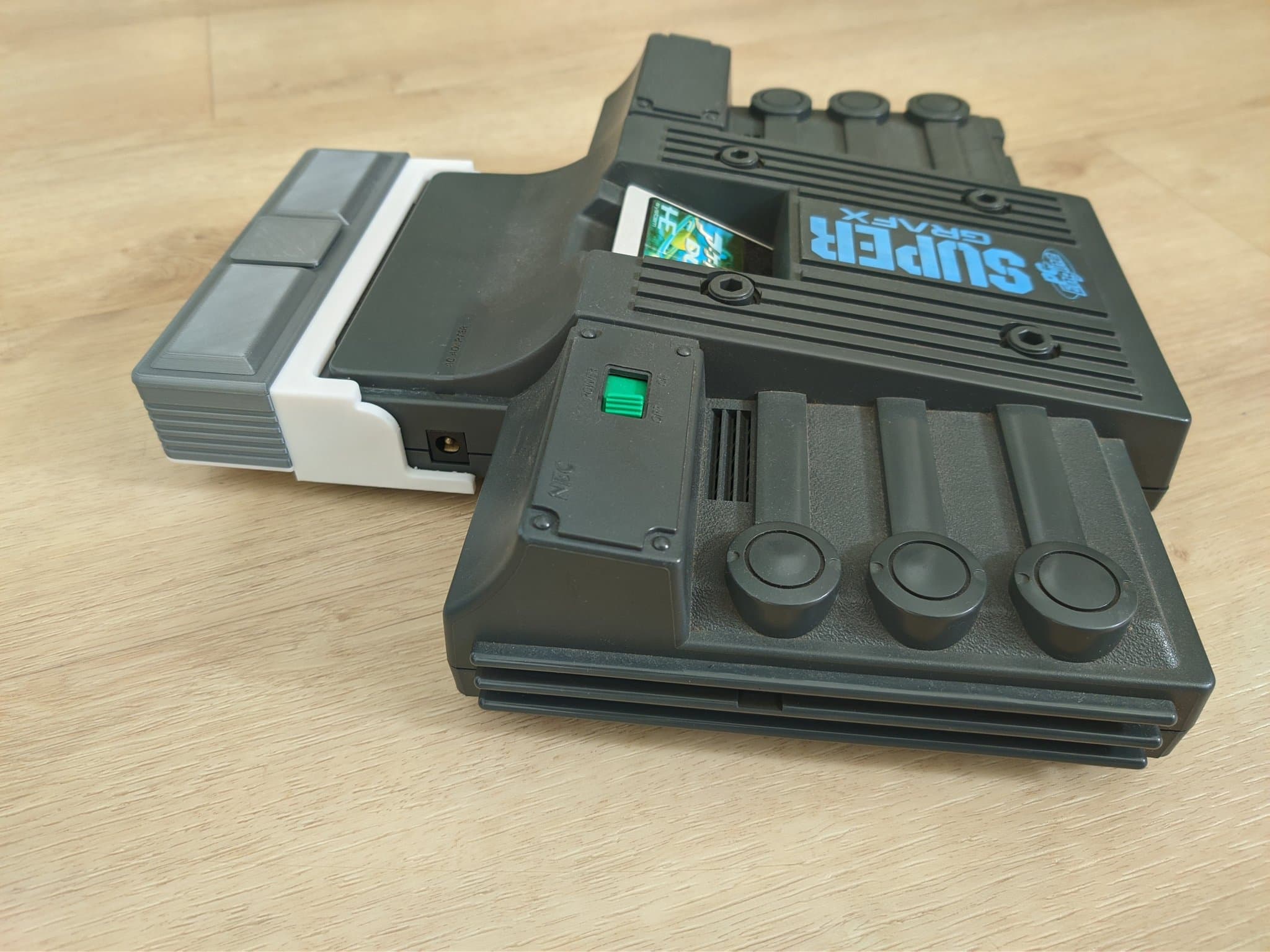 Refurbished NEC PC Engine SuperGrafx with the EDFX in its mini-IFU shell connected and the green power indicator lit, on a wooden surface; the raised details on the shell are part of the SuperGrafx styling, not control knobs.