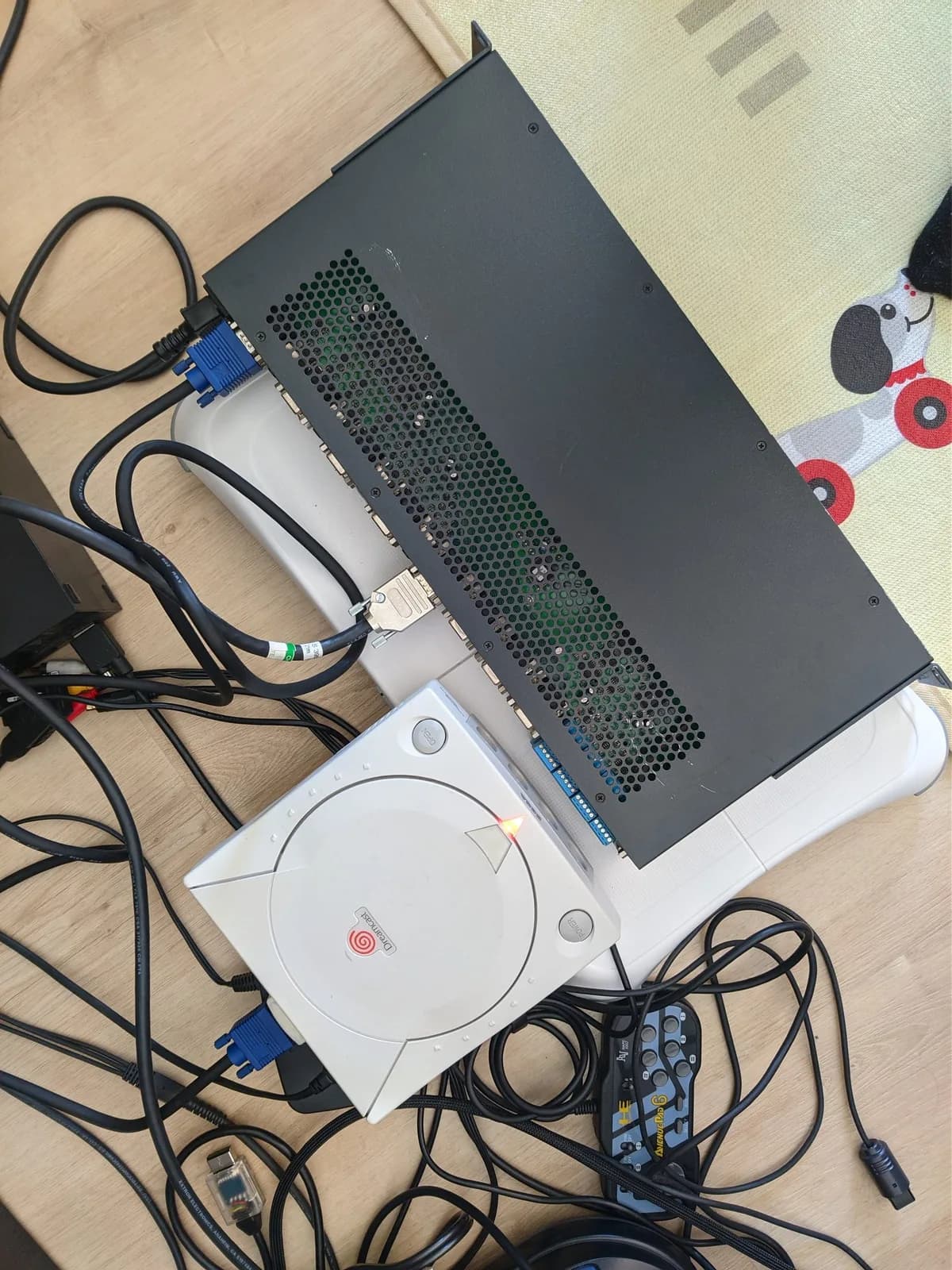 Overhead shot: a white Sega Dreamcast (powered on) behind the black Extron MVX switcher, connected through a Jeff Chen DC2VGA dongle amid tangled cables on a wooden desk.