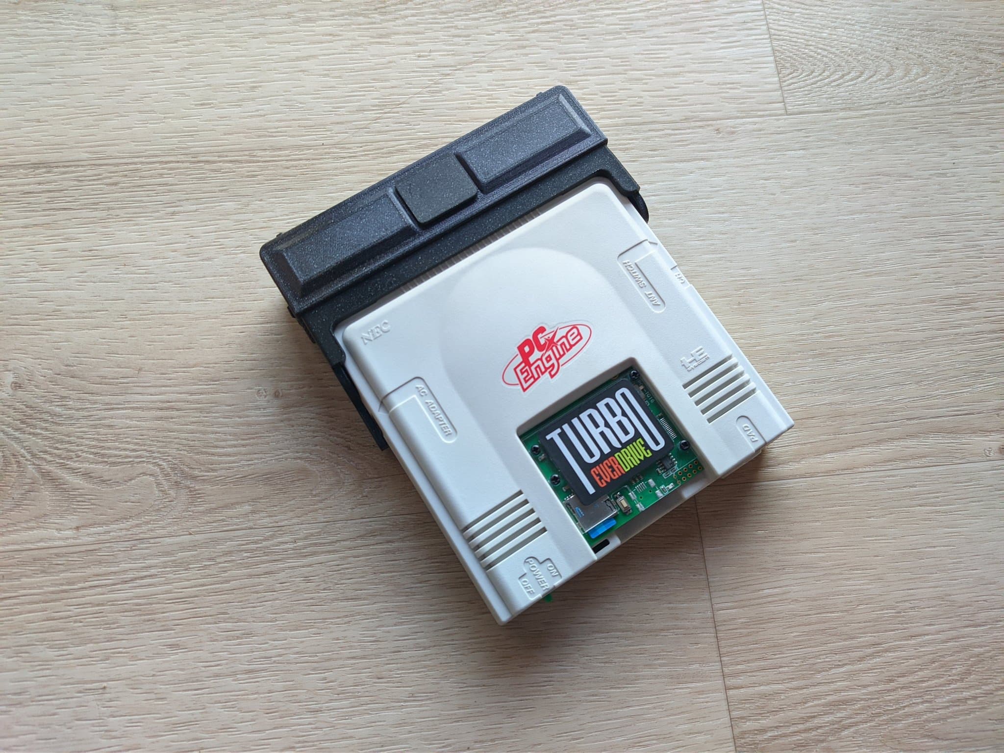 The PC Engine with the AV adapter connected and a Turbo Everdrive v2 (the green cartridge labelled TURBO) inserted in the HuCard slot, on a wooden surface.